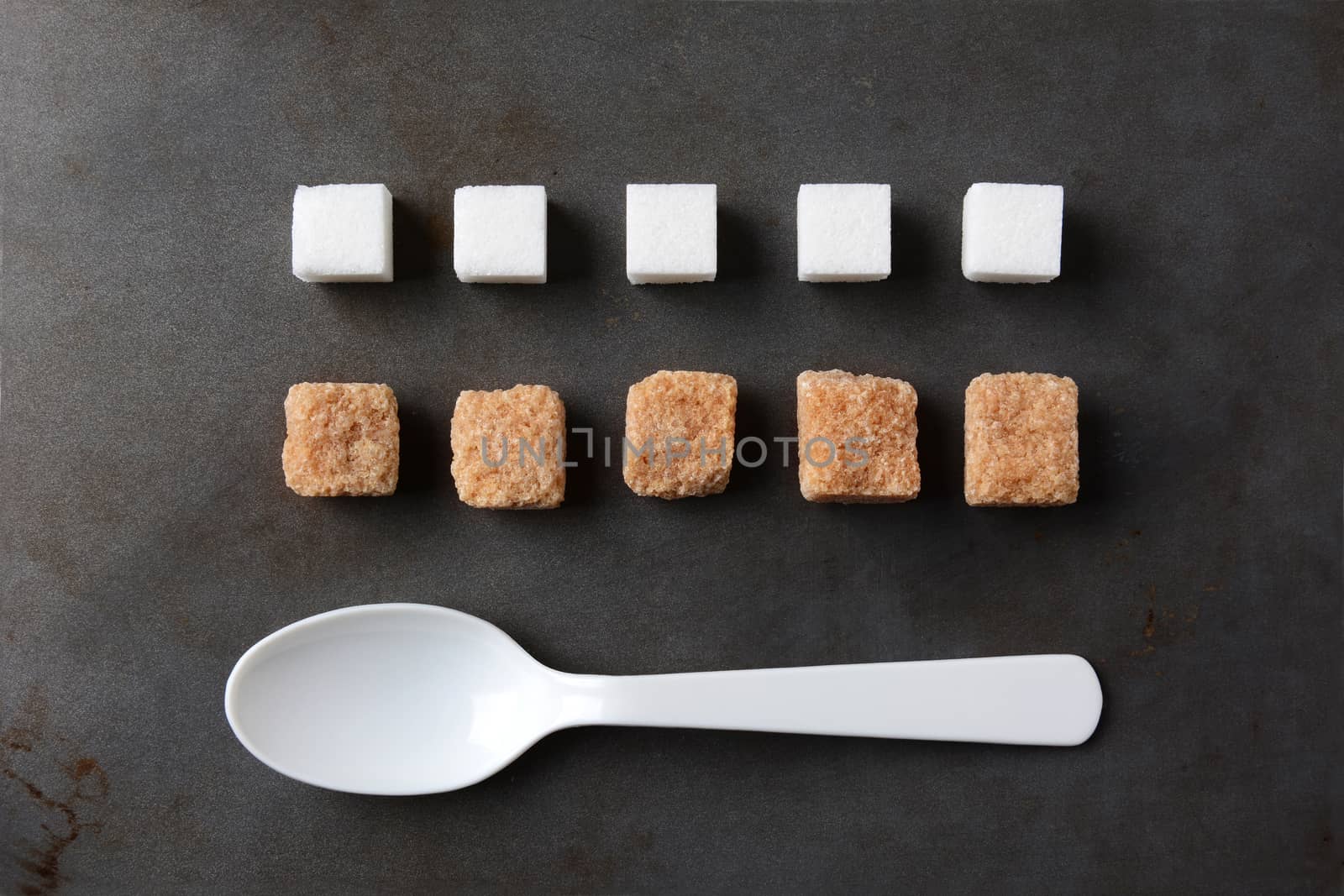 High angle view of white and brown sugar cubes and a white plastic spoon on a metal baking sheet. Five cubes of both the white and brown lumps line up in rows. Horizontal format.