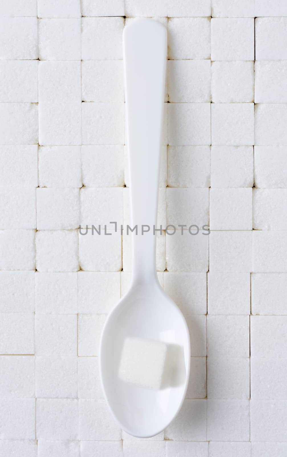 A white plastic spoon with a single sugar cube on a background of closely stacked white sugar cubes.