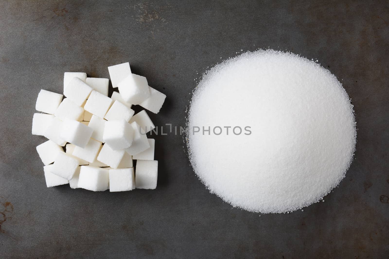 Overhead view of a pile of granulated white sugar and a mound of sugar cubes, on a used baking sheet.