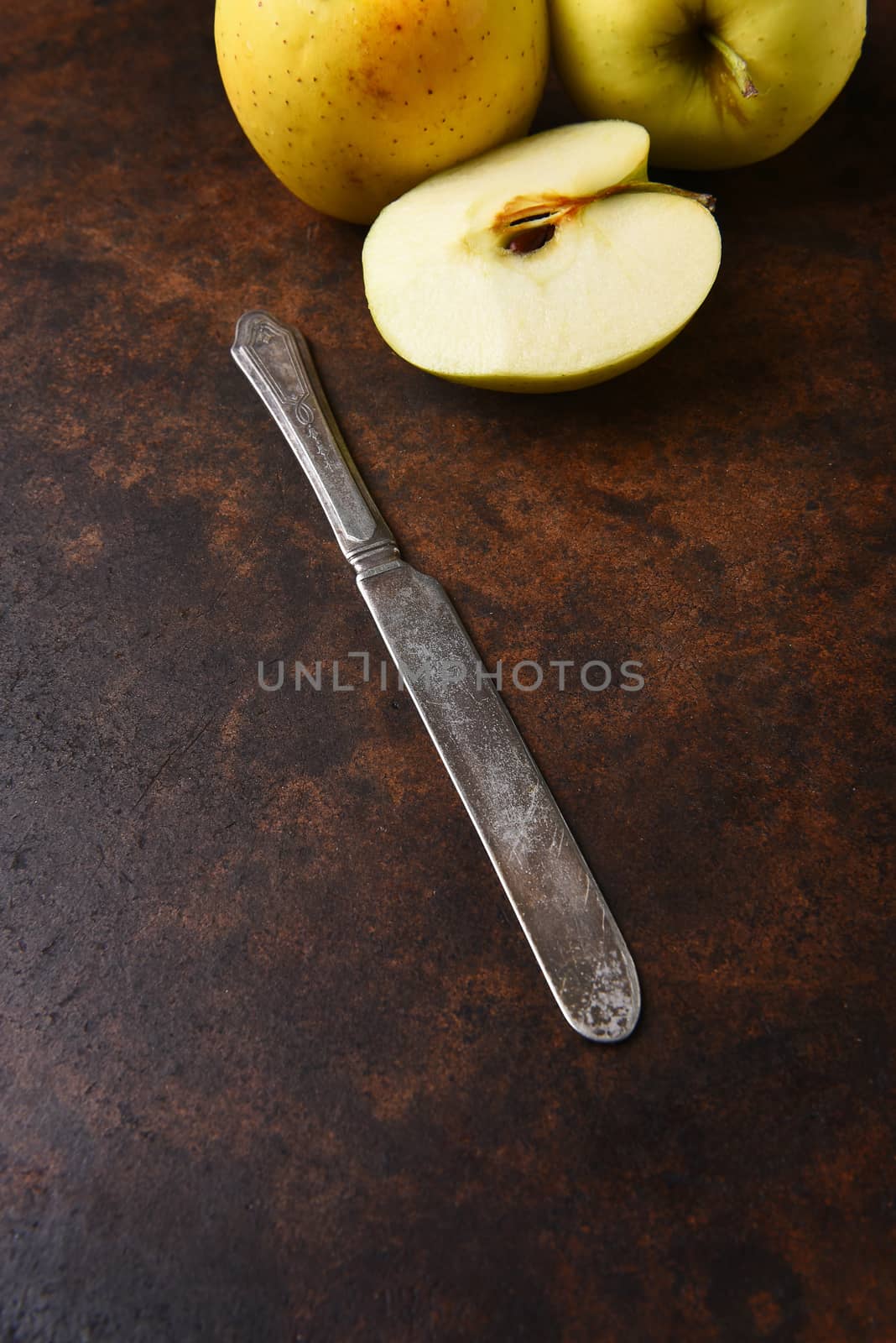 Closeup of an old knife with golden delicious apples in the background. Vertical format on a dark mottled surface.