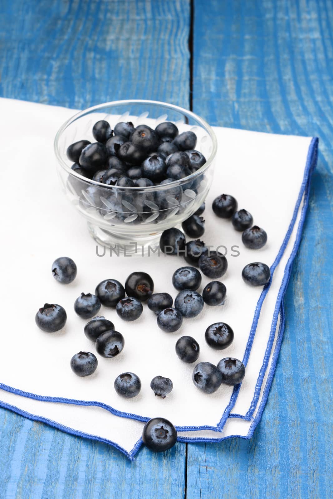 Closeup of a group of blueberries scattered on a napkin with a glass bowl filled with more berries. Vertical format on a rustic farmhouse style painted table, with shallow depth of field.