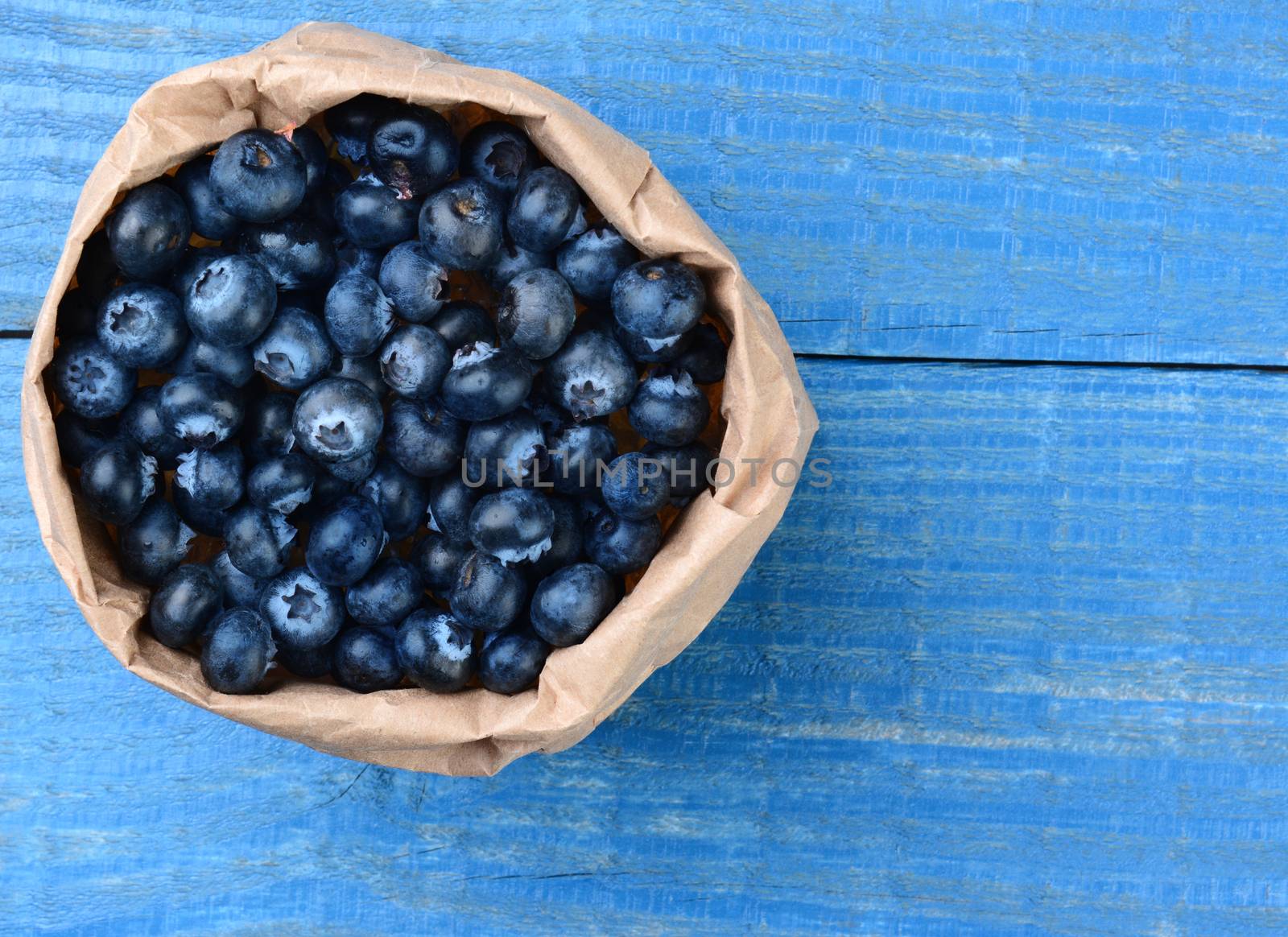 A brown paper bag full of fresh picked blueberries. The bag is on a blue wooden table.