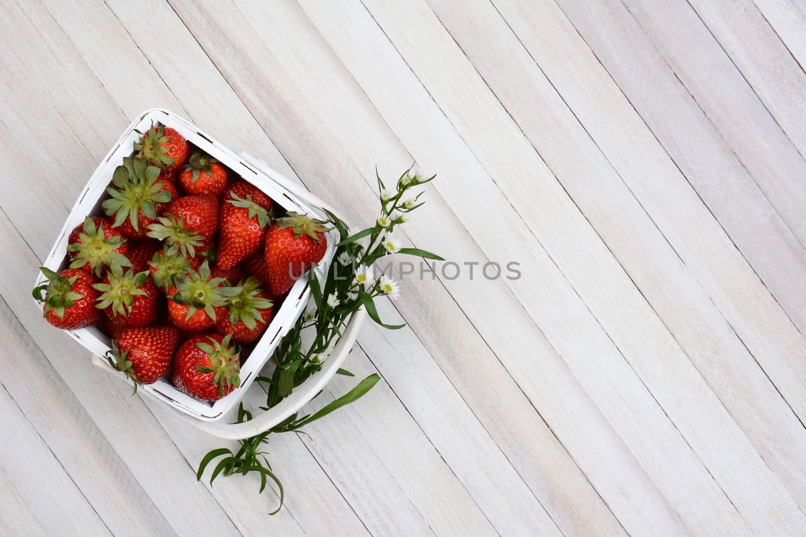 Overhead view of a basket of fresh picked strawberries on a white rustic wood table. A sprig of flowers is stuck between the handle and the basket. Horizontal format with copy space.