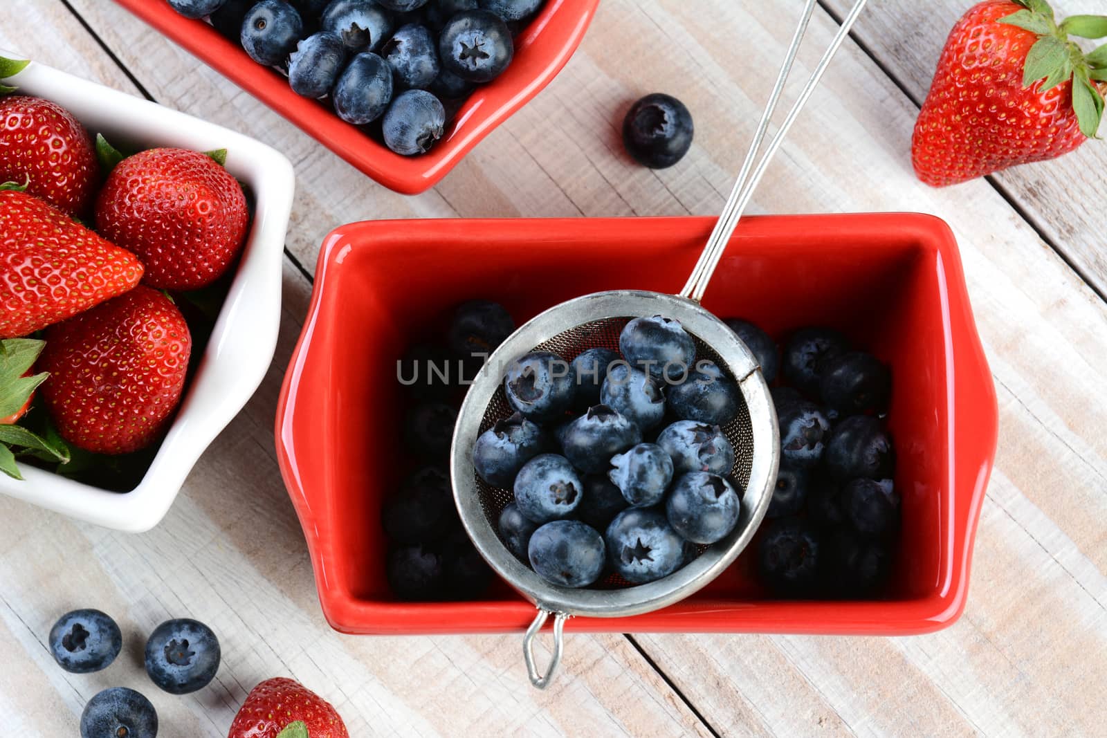 High angle shot of fresh picked berries in red and white bowls and a strainer. Strawberries and Blueberries in horizontal format on rustic wood farmhouse style table.