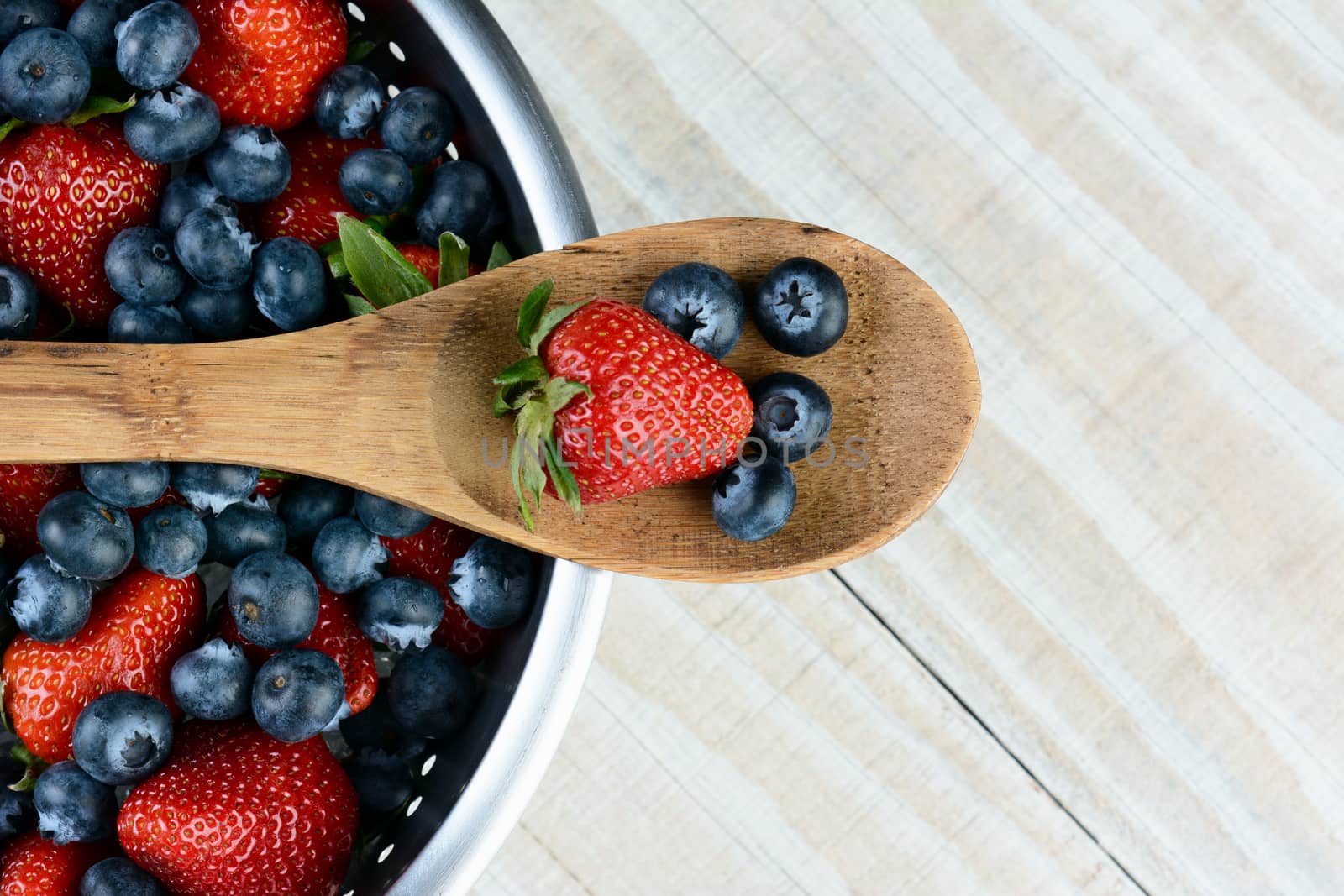 High angel photo of a Strawberry and Blueberries on a wooden spoon laying across a colander full of berries on a rustic wooden farmhouse style table. Horizontal format with copy space.