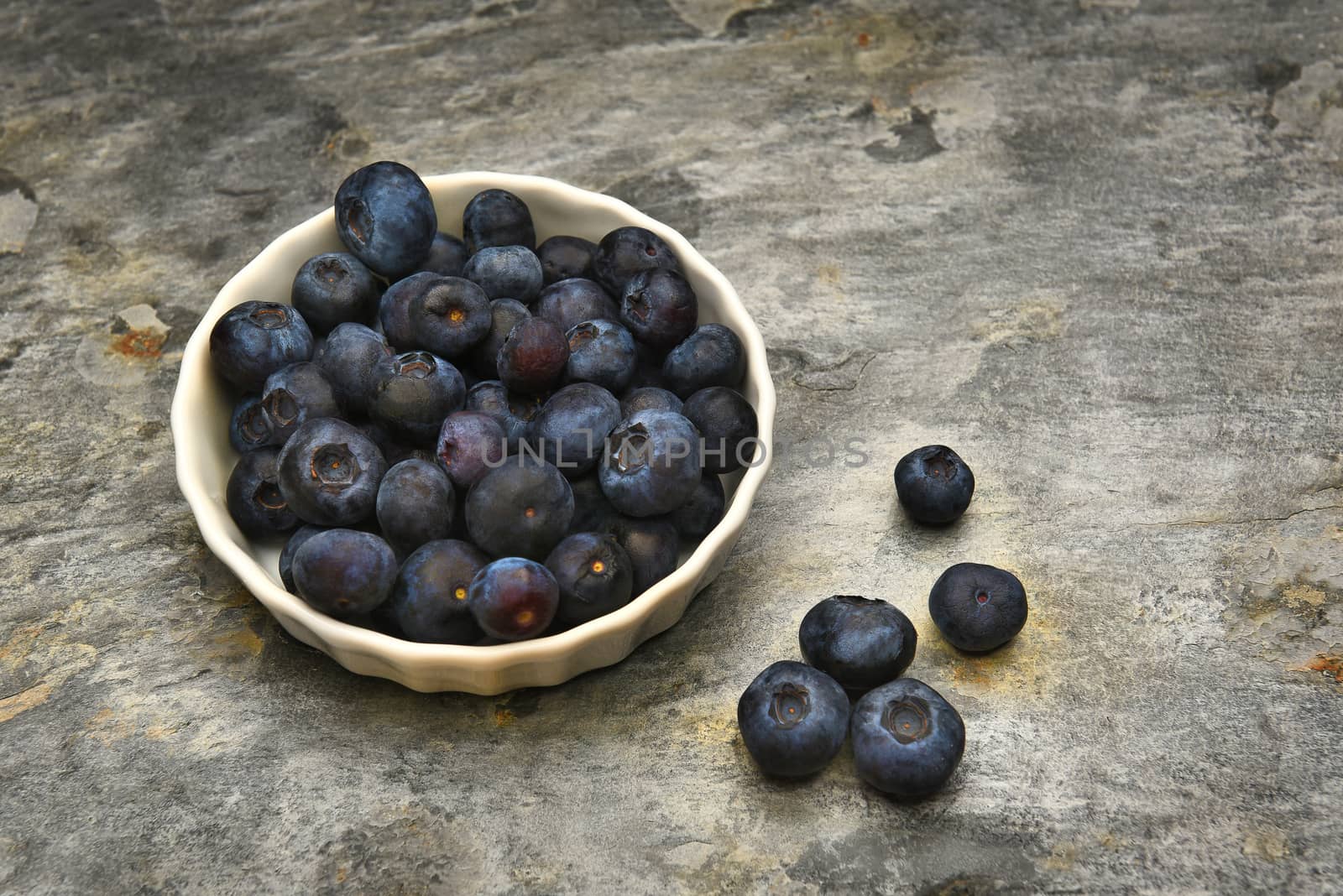 A ramekin filled with fresh ripe blueberries on a slate table top, with loose berries scattered on the table.