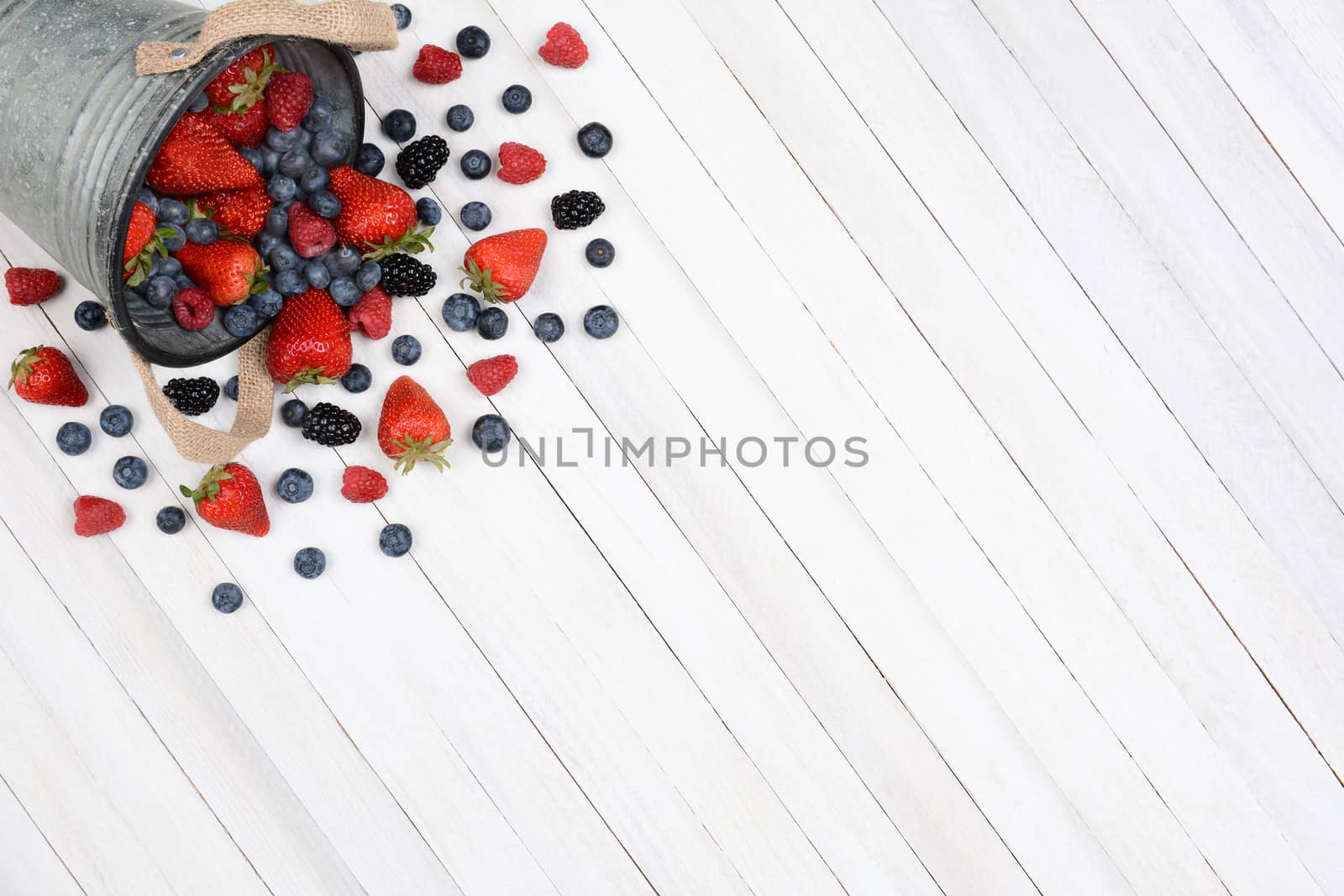 A bucket of berries spilling onto a rustic white kitchen table. High angle shot with the pail in the upper left corner leaving room for your copy.