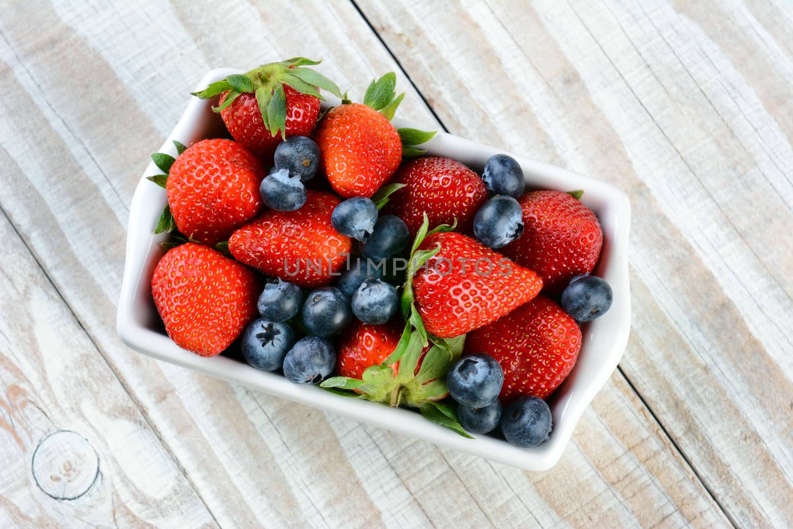 A rectangular bowl full of berries. Blueberries and strawberries on a rustic wooden kitchen table.