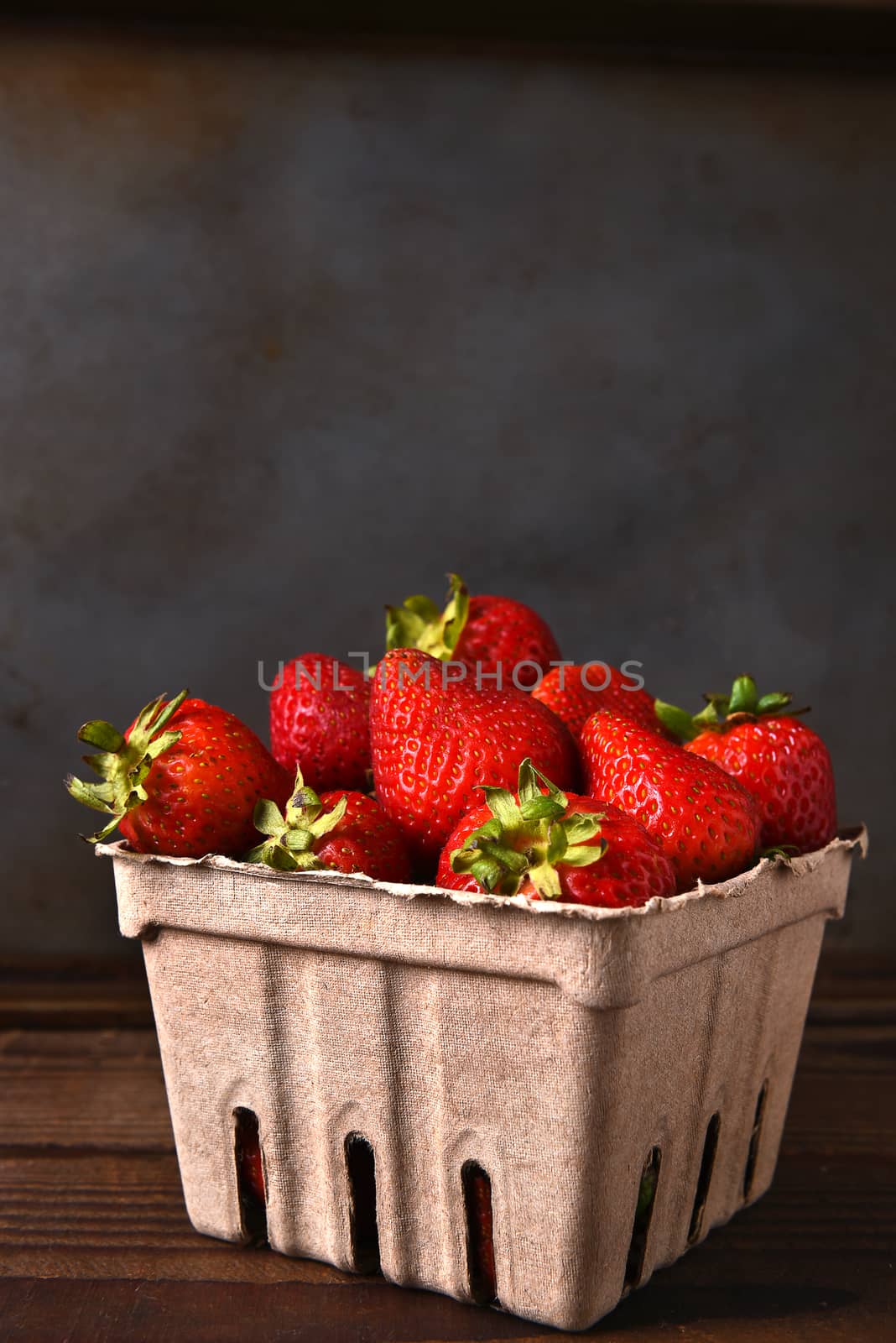 Still life of a cardboard produce container full of fresh picked strawberries on a wood table. Vertical format with copy space.
