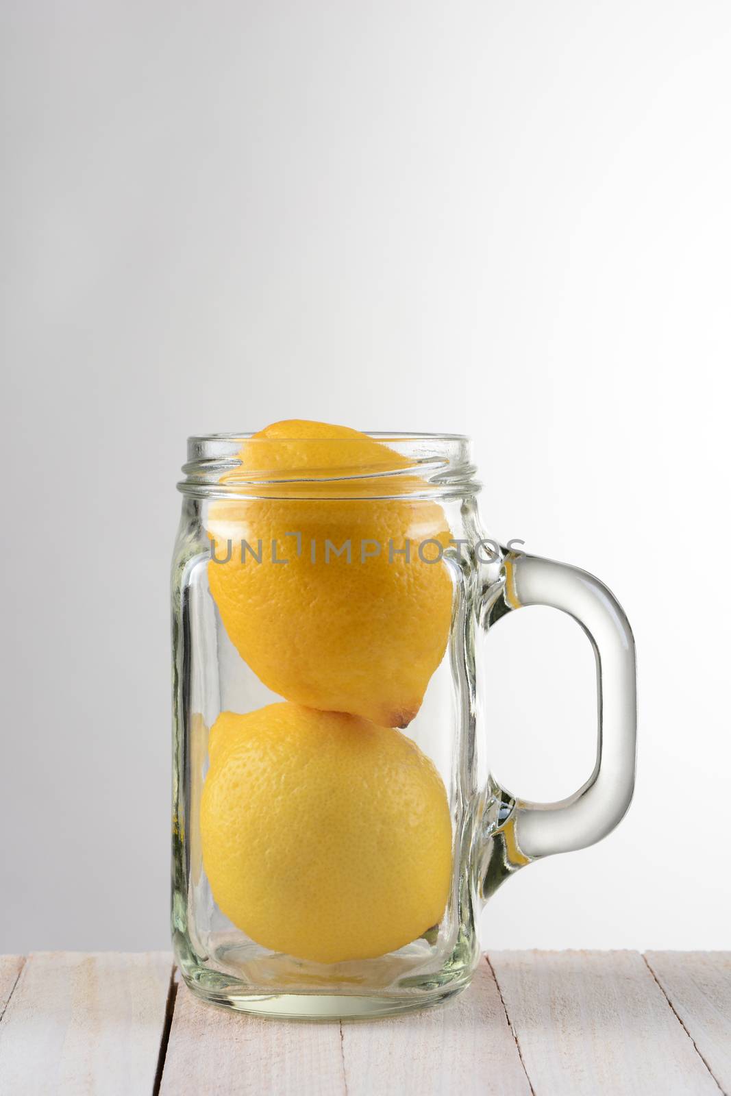 A still life with two lemons in a mason style jar with handle. The glass is on a rustic white wood table against a light to dark gray background with copy space.