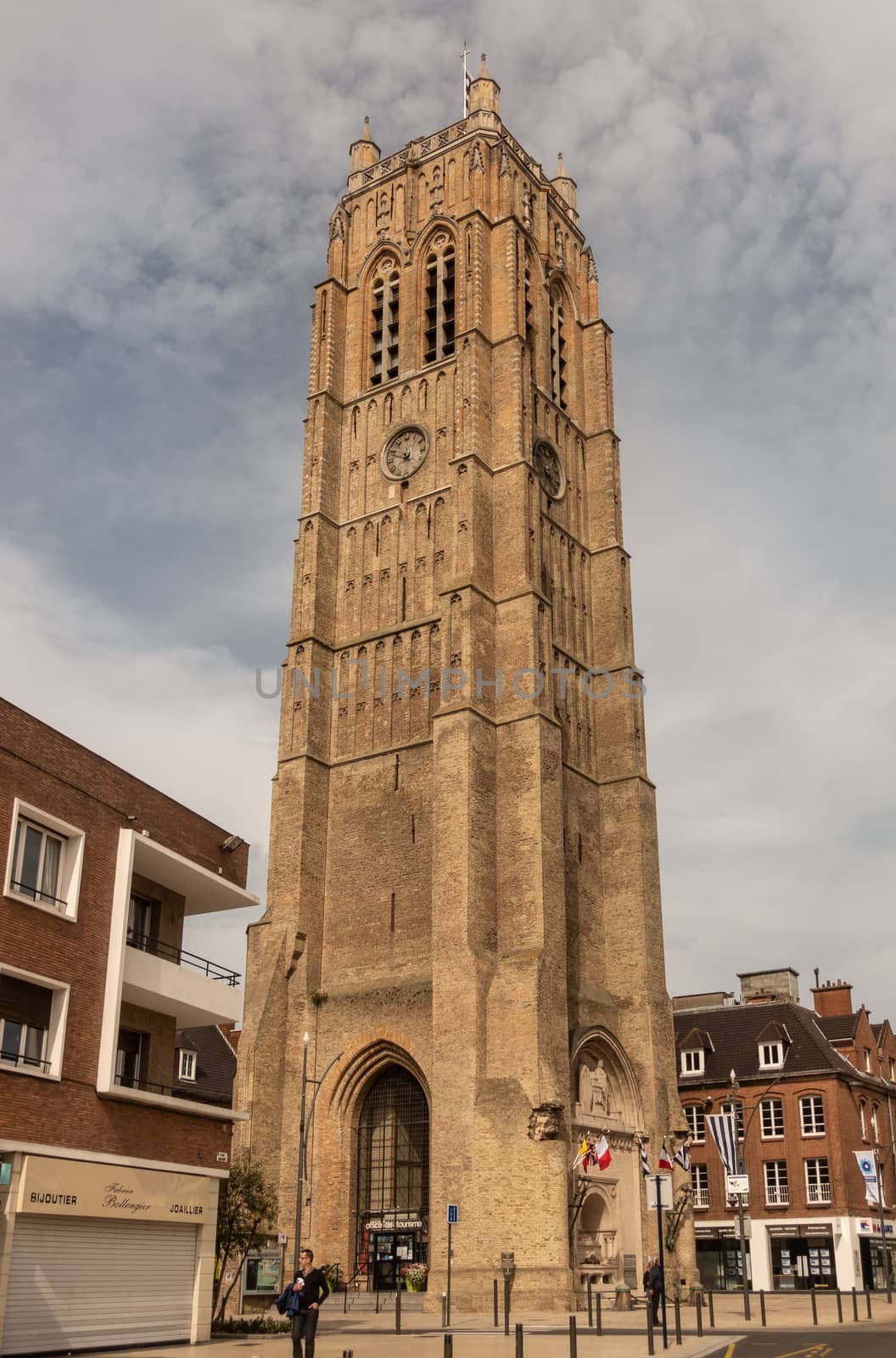 Dunkerque, France - September 16, 2018: the brown brick Belfry of Dunkirk towers over houses under cloudy blue sky. Street scene with people.