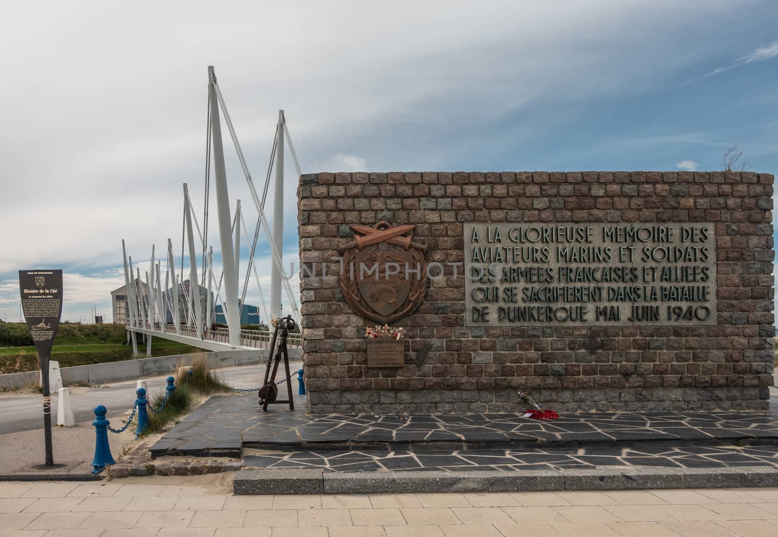 Dunkerque, France - September 16, 2018: Brown stone war memorial commemorating the Battle for Dunkirk in world war 2 near the beach. Pedestrial suspension bridge under blue-white sky.