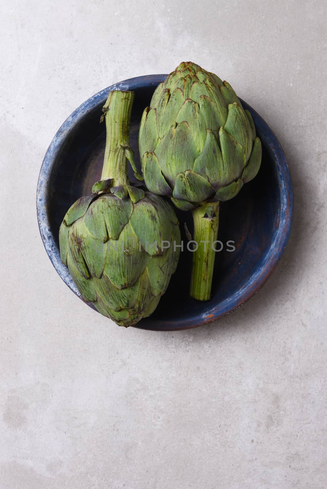 Two artichokes in a blue bowl on a mottled gray background with copy space.