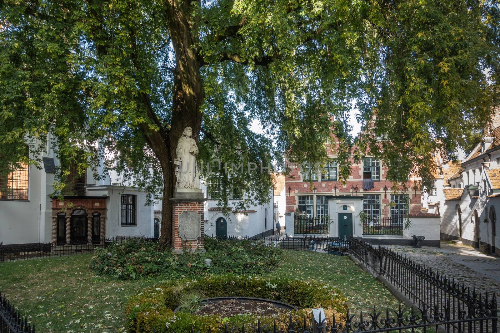 Kortrijk, Flanders, Belgium - September 17, 2018: White Johanna of Constantinople statue under green tree in beguinage courtyard. Small houses around.