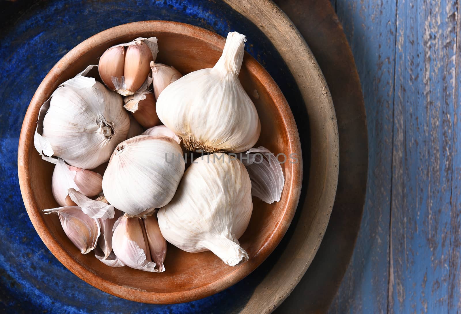 HIgh angle view of a bowl of garlic bulbs and cloves in a blow, on a rustic blue table. 