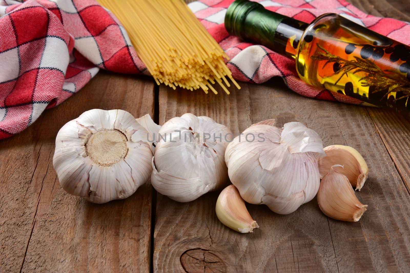 Italian cooking still life on a rustic wood table. Garlic cloves in the foreground with olive oil dried spaghetti and red and white checked napkin. Shallow depth of field.