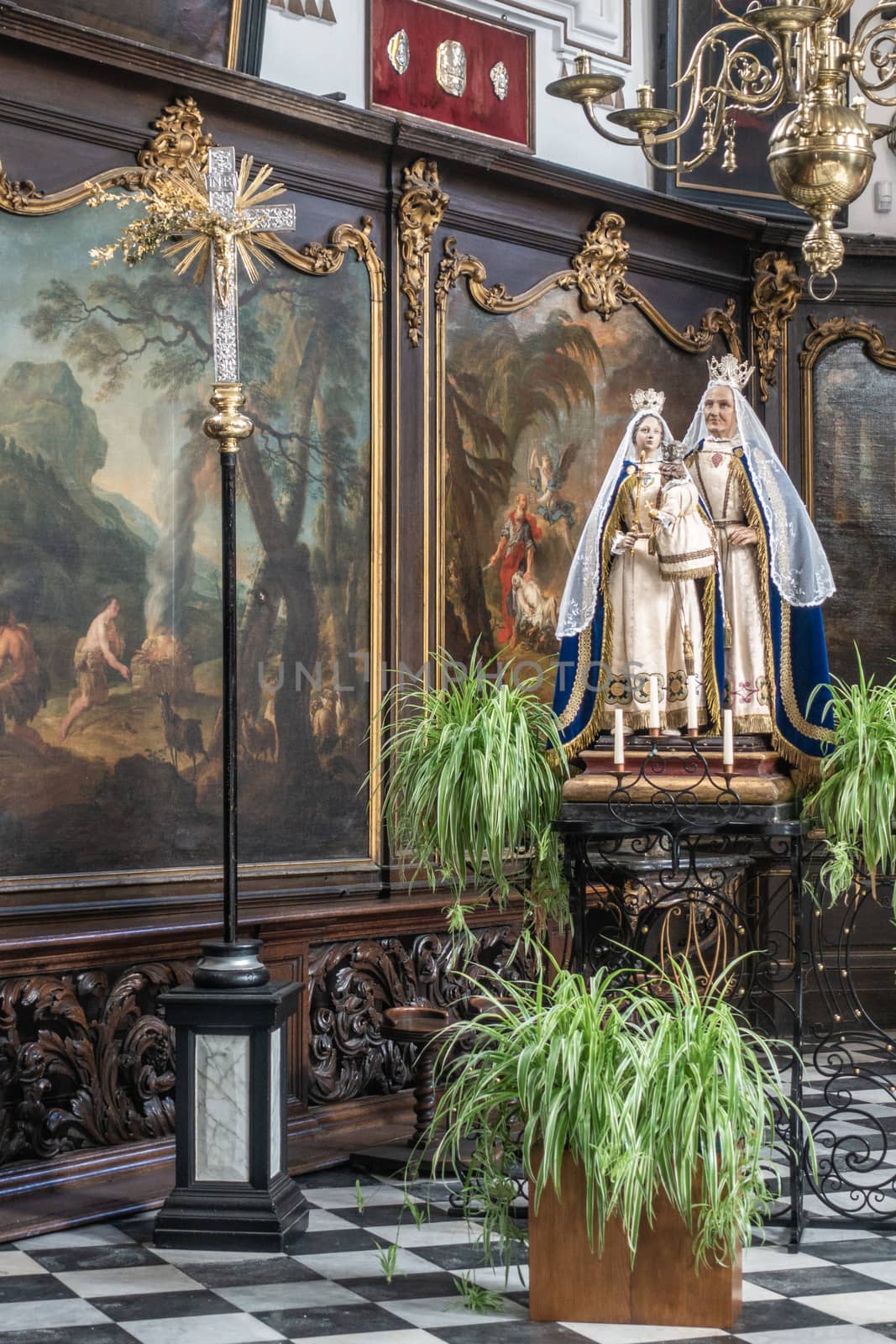 Brugge, Flanders, Belgium - September 19, 2018: Wall covered in paintings with gold decorations behind statue of Saint Anna and the Madonna in Saint Anna Church in Bruges. Some green plants up front.