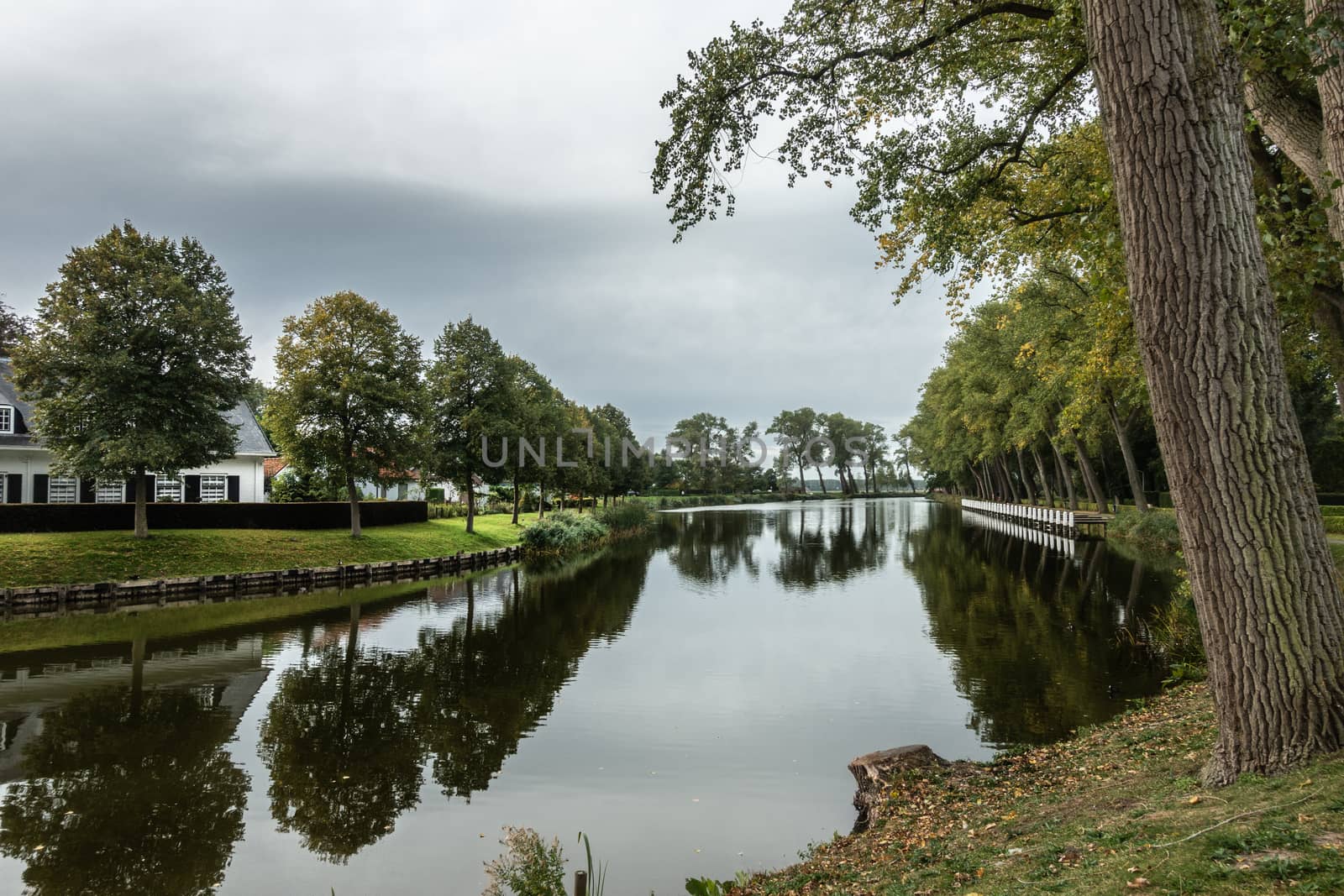Sluis, Zeeland, Netherlands - September 22, 2018: Gray sky and green bordering trees reflected in quiet water of dead ending canal Bruges-Sluis in Sluis. White house.