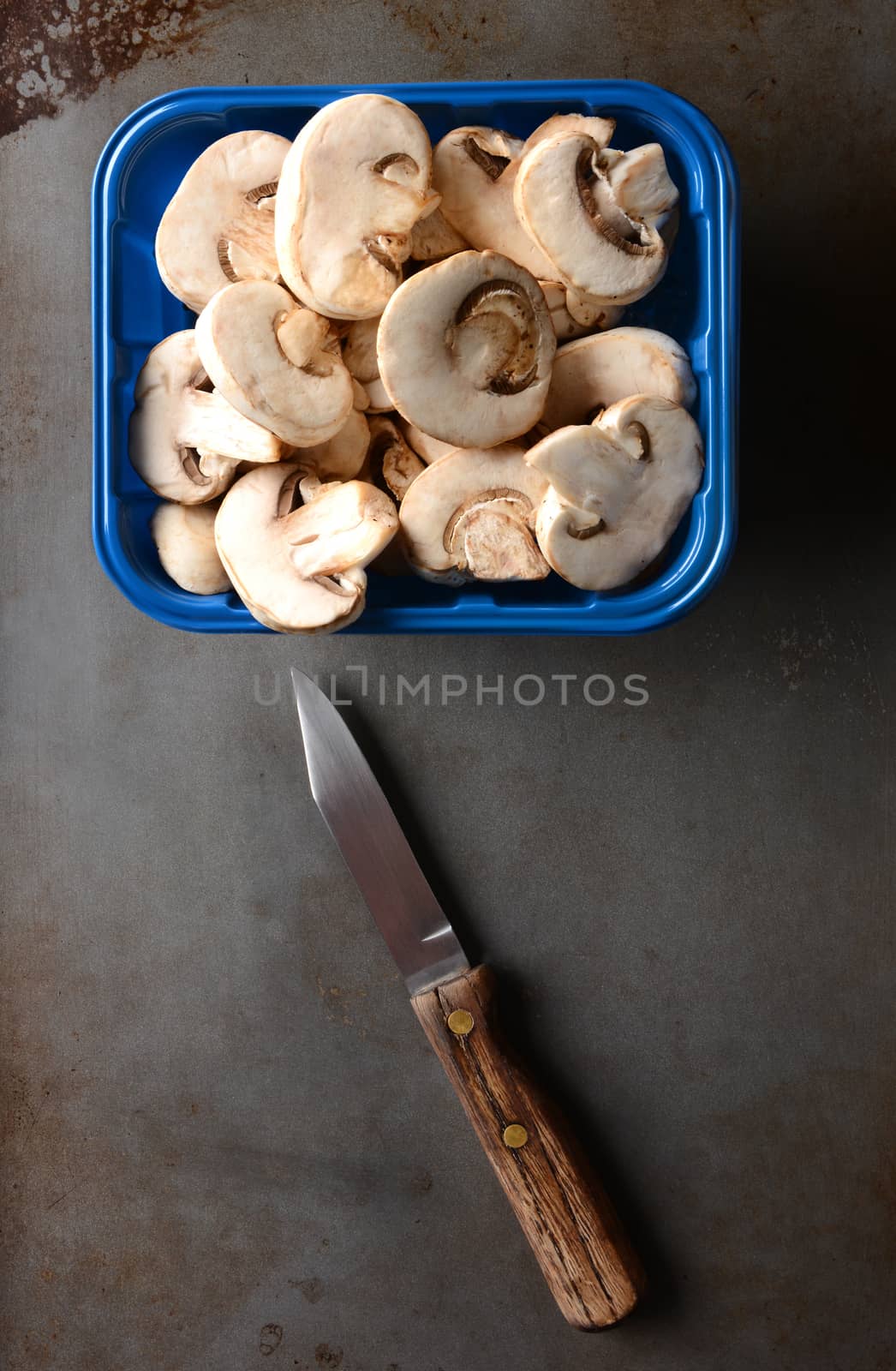 A blue container of sliced mushrooms on a metal baking sheet with a knife. Overhead shot in vertical format.