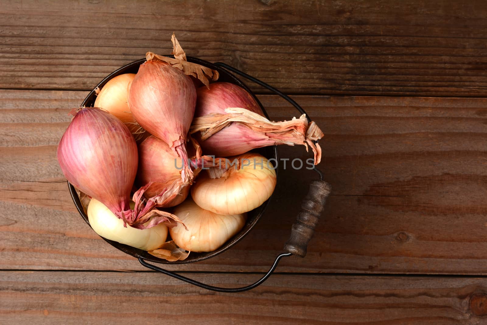 Overhead view of onions and shallots in a bucket on a rustic wooden table. Horizontal format.