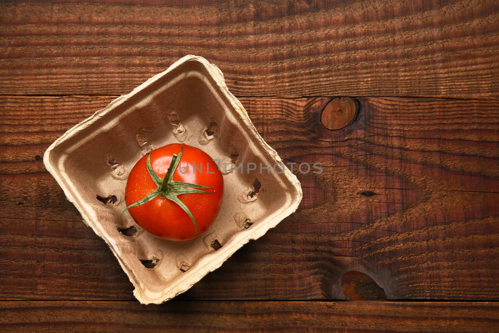 High angle view of a single tomato in a cardboard produce container on a dark wood surface.