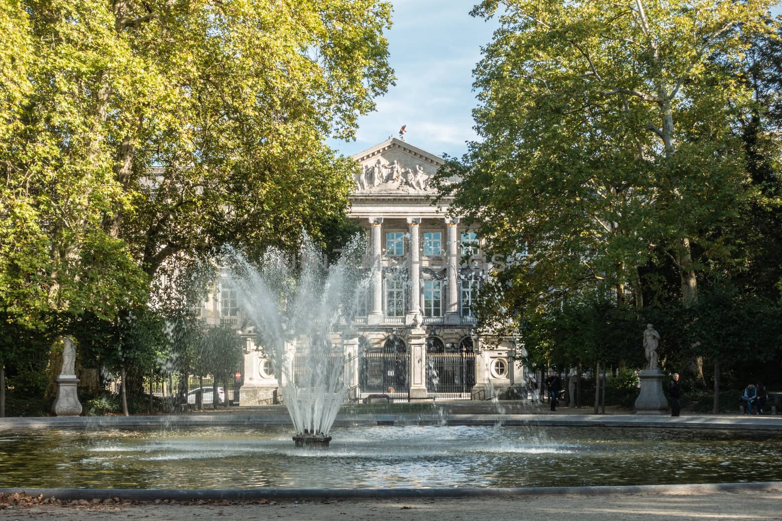 Brussels, Belgium - September 26, 2018: Fountain in Park of Brussels partly obscures facade of Belgian Parliament. Green foliage, statues and people on bench.
