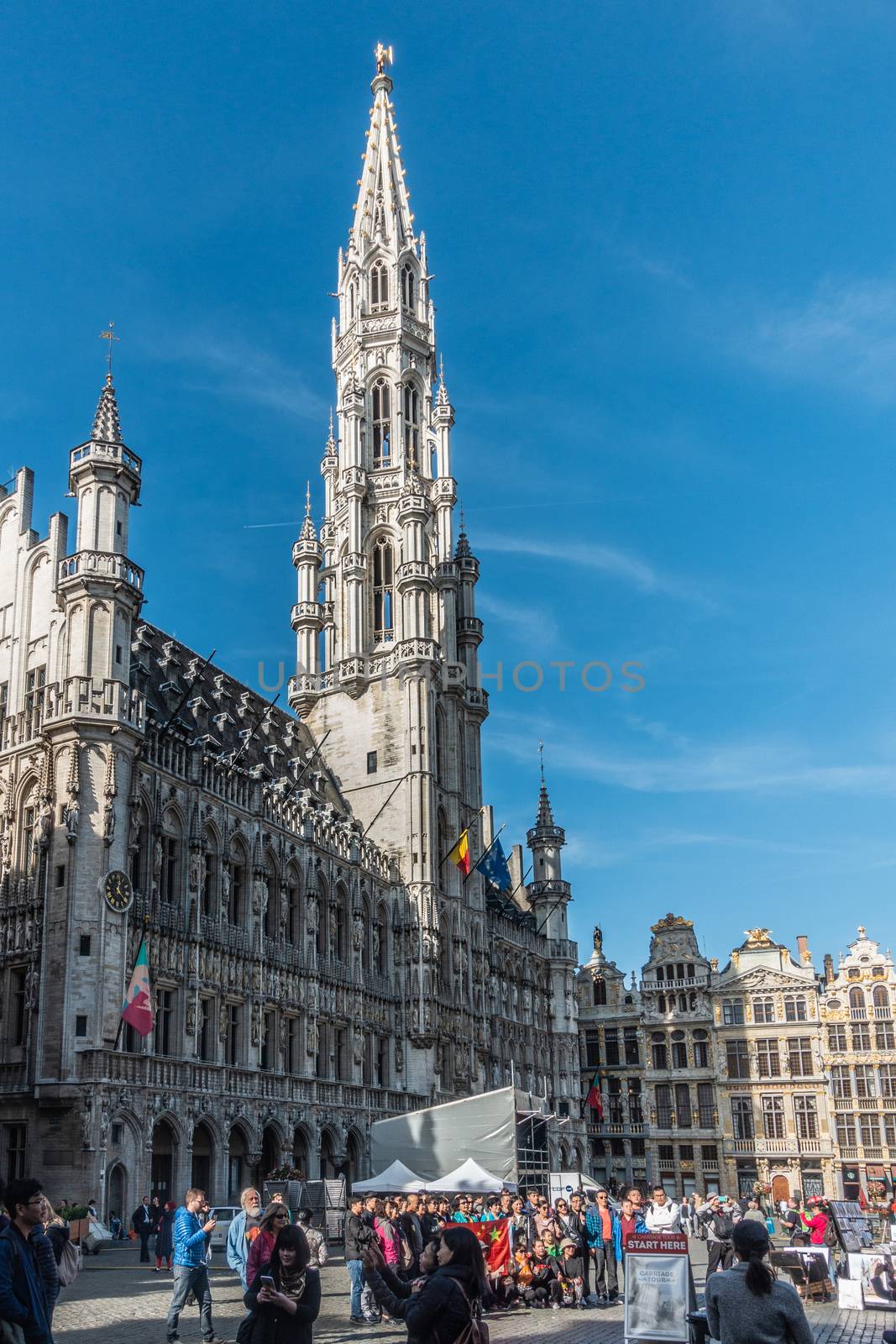 Brussels, Belgium - September 26, 2018: Town hall with its gray stone spire on the Grand Place, Grote Markt, with plenty of people and facades of other houses with golden statues. Blue sky.