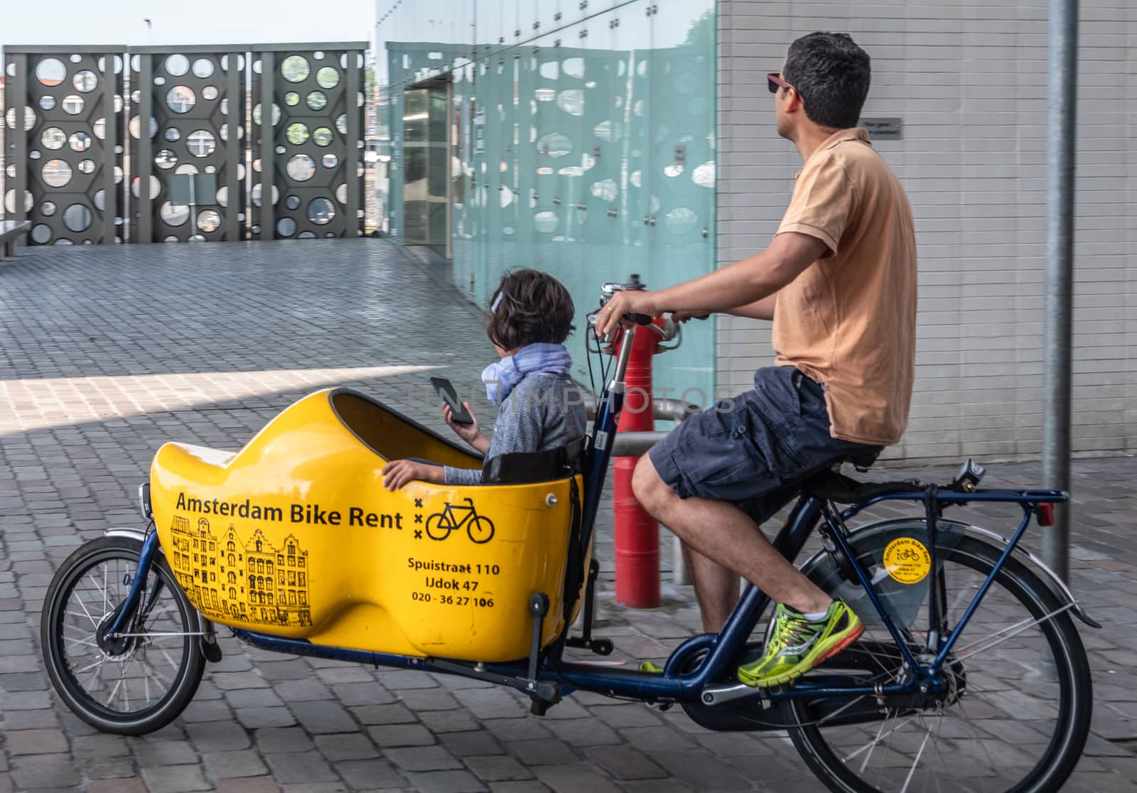 Amsterdam, the Netherlands - June 30, 2019: Closeup of bakfiets, front-trunk bike to transport a load, a child here. Man pedaling as seen on IJdok. Trunk in shape of yellow clog.