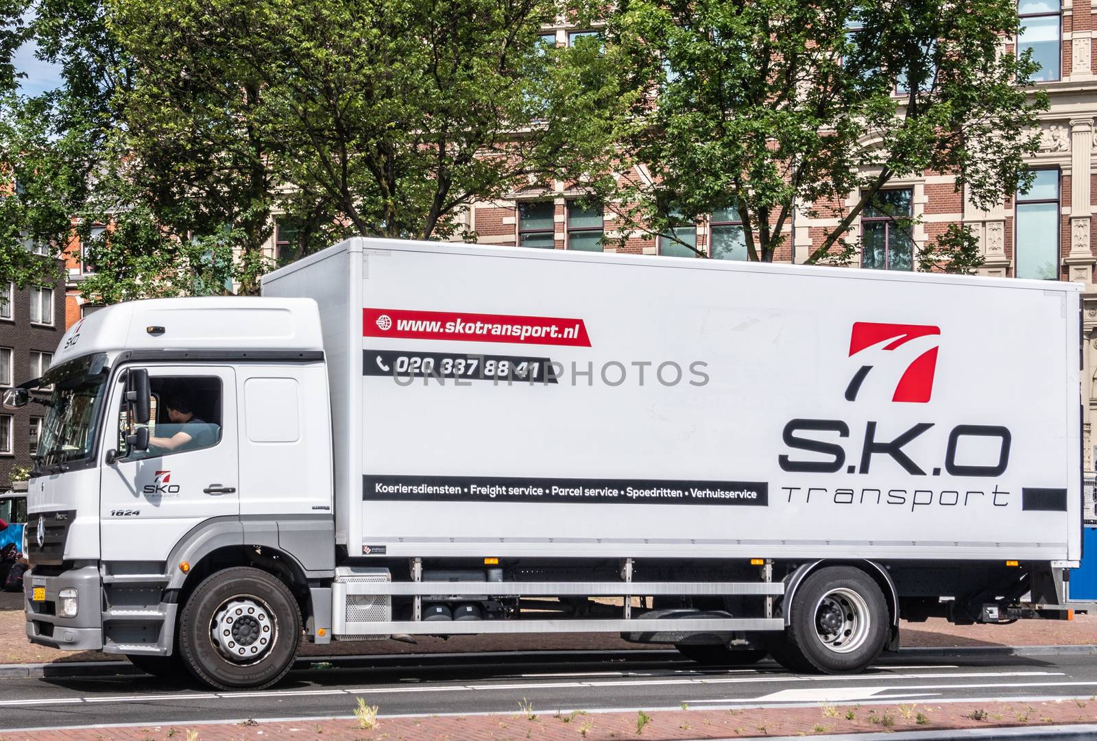 Amsterdam, the Netherlands - June 30, 2019: Large white transport truck with black and red lettering for S.K.O. company. Green foliage above. Brown-white facades in back.