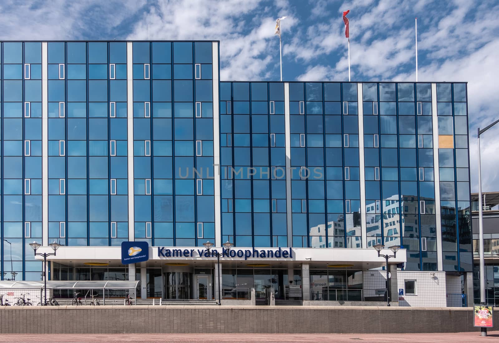 Amsterdam, the Netherlands - June 30, 2019: Modern glass facade building of Chamber of Commerce, Kamer van Koophandel, along De Ruijterkade opposite Ijdok, with flags and blue white sky.