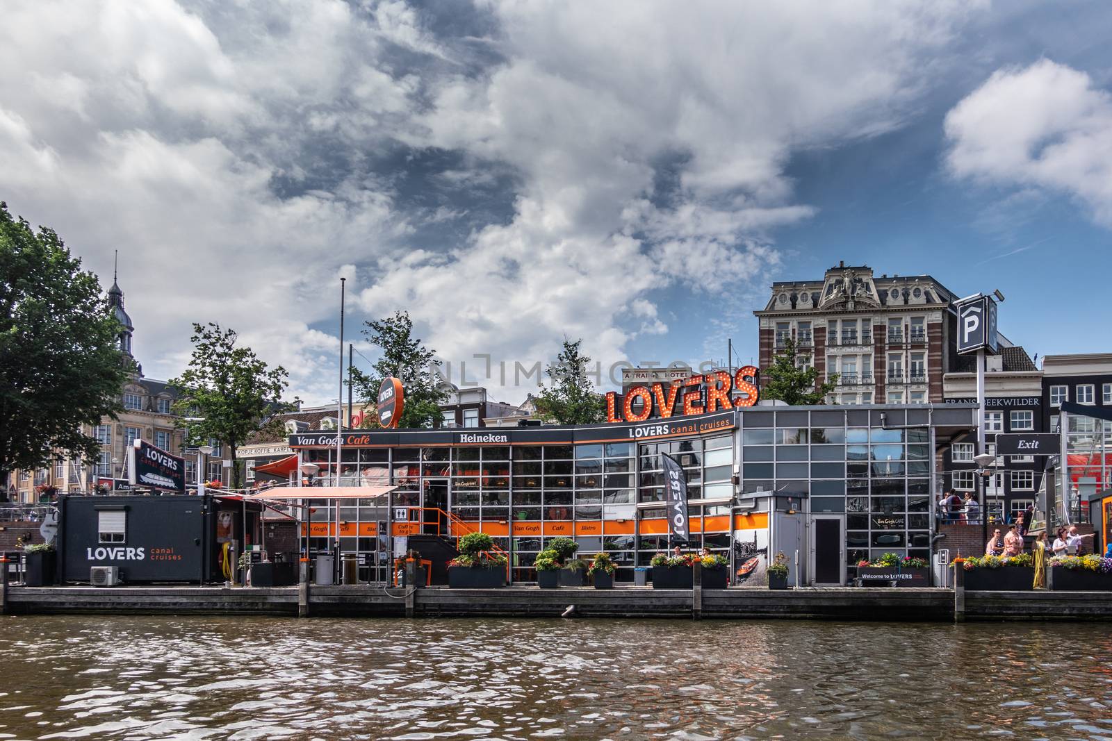 Amsterdam, the Netherlands - June 30, 2019: gray white cloudscape with blue patches above Orange decorated Lovers Canal cruises office along canal. Some green foliage and houses in back.
