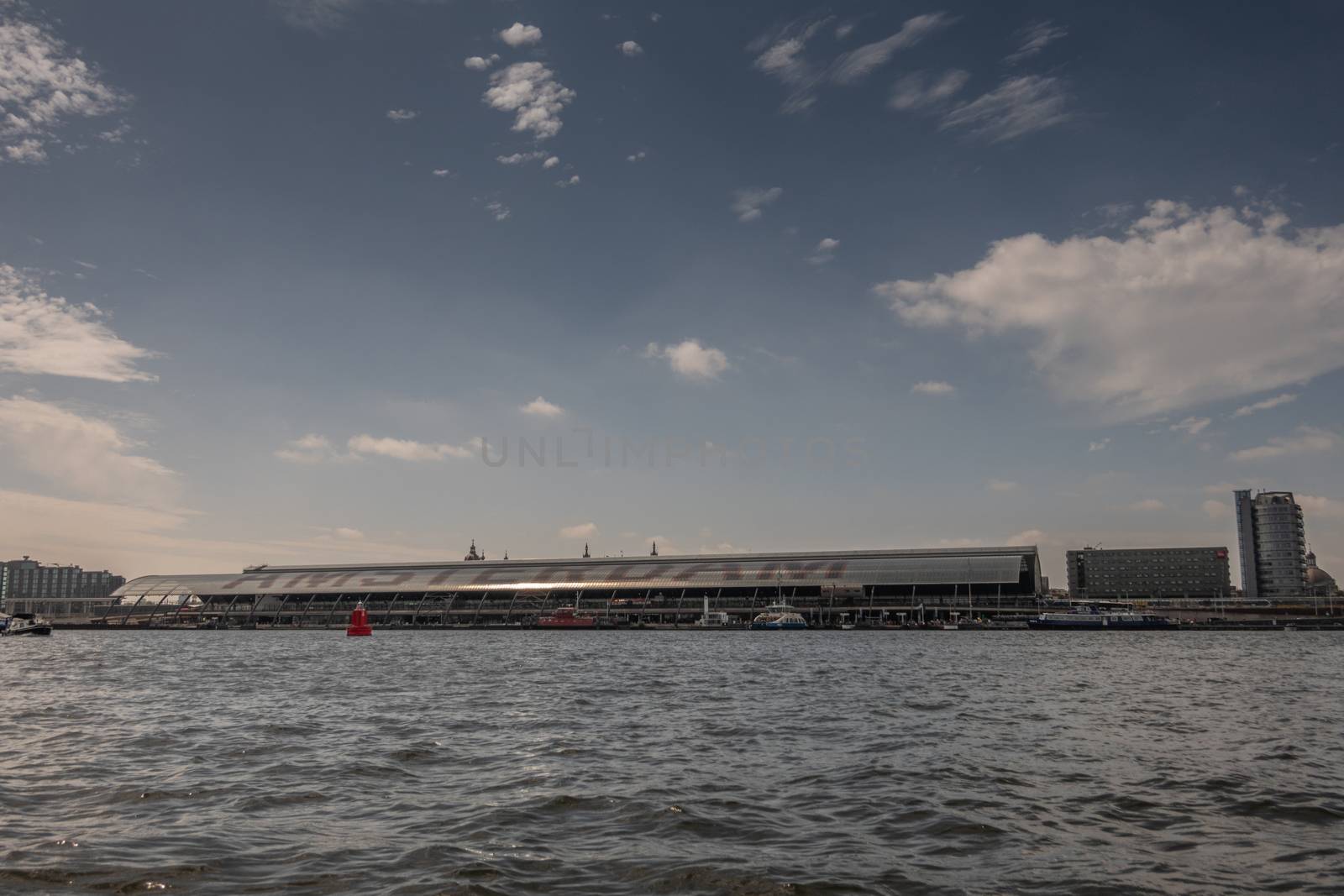 Amsterdam, the Netherlands - June 30, 2019: Terminal hall of Centraal Railway Station seen from IJ water under blue sky with white clouds. Other buildings at the edges.