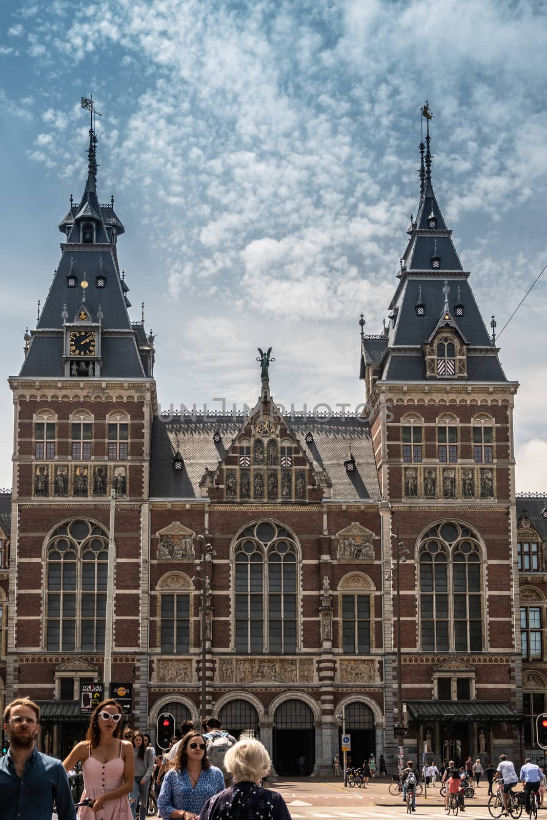Amsterdam, the Netherlands - June 30, 2019: Monumental entrance facade with towers in beige and red bricks. Statues and frescoes. Under blue-whtie cloudscape. People in front.