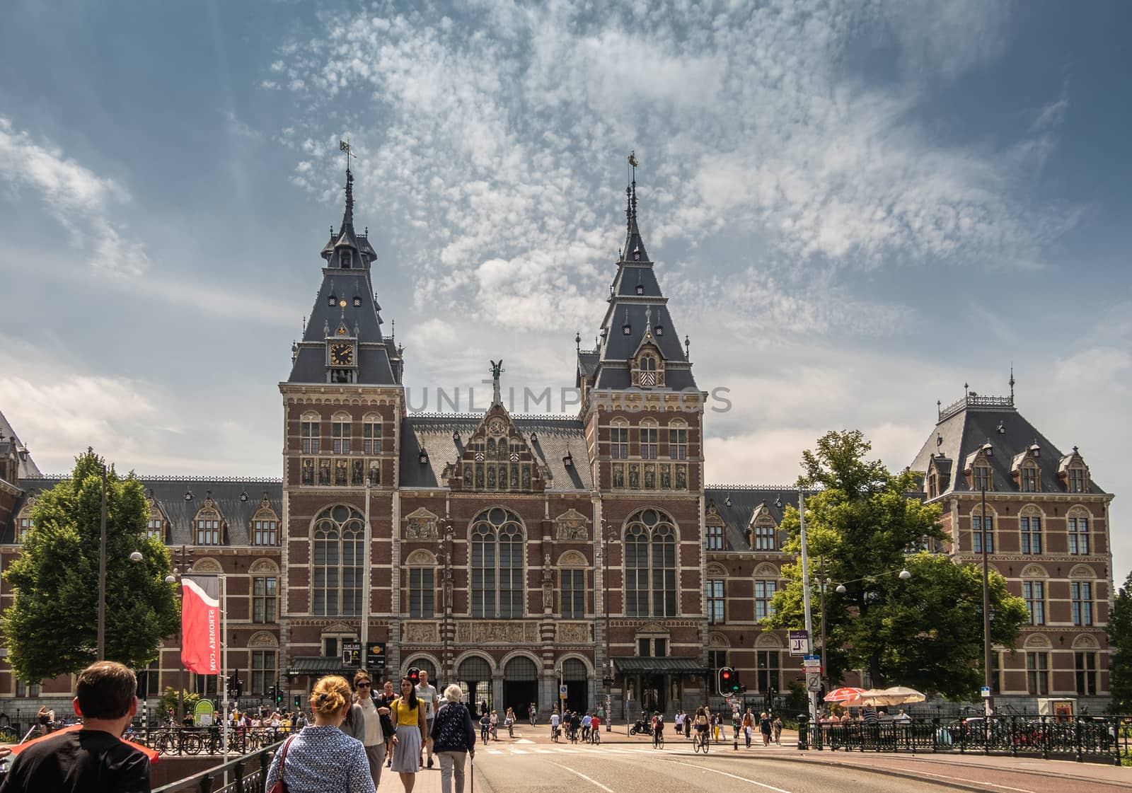 Amsterdam, the Netherlands - June 30, 2019: Wide shot of Monumental facade with towers in beige and red bricks. Statues and frescoes. Under blue-whtie cloudscape. People in front.