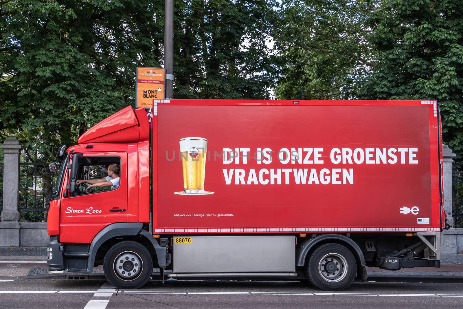 Amsterdam, the Netherlands - July 1, 2019: Red Amstel beer delivery truck although it claims to be the greenest truck. Driver active. Image of full Amstel bier glass. Green foliage.