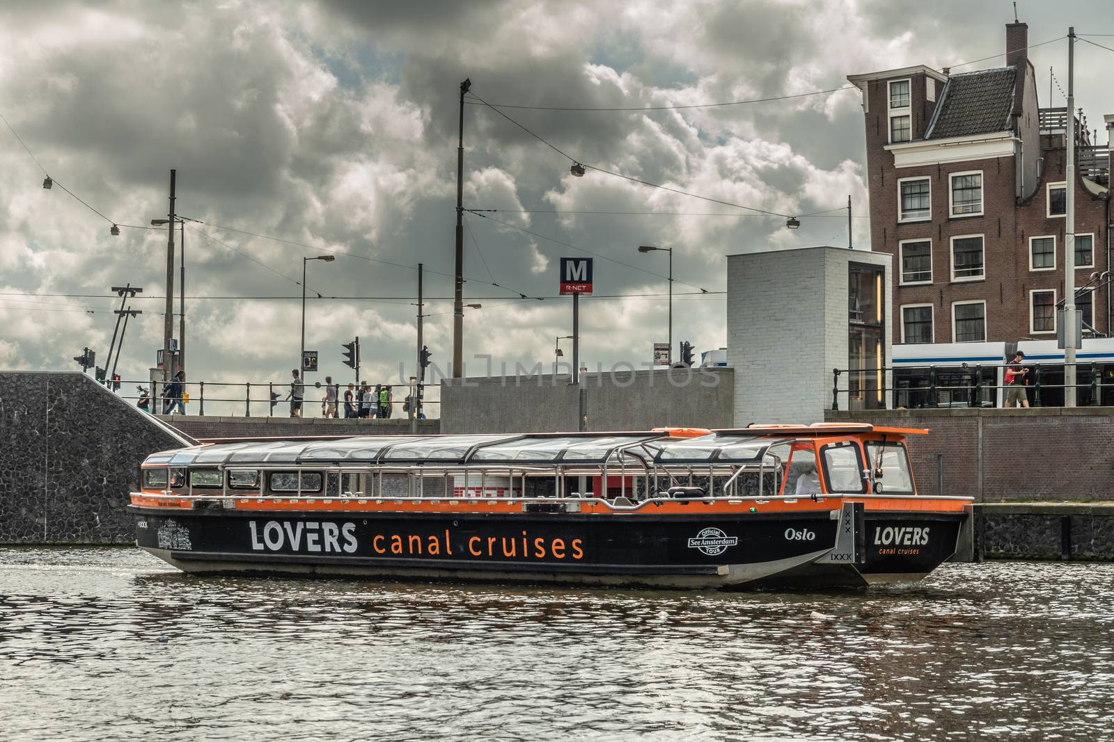 Amsterdam, the Netherlands - July 1, 2019: Lovers Canal Cruises boat on the move in canal under heavy cloudscape on top of dark water. Street with houses and people in back.