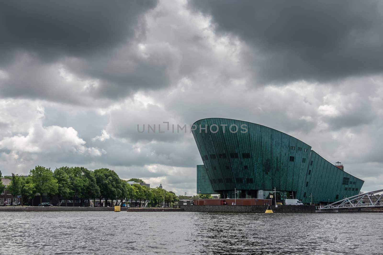 Amsterdam, the Netherlands - July 1, 2019: Modern boat hull building housing the NEMO science museum on border of Oosterdok in black water under havey rainy cloudscape.