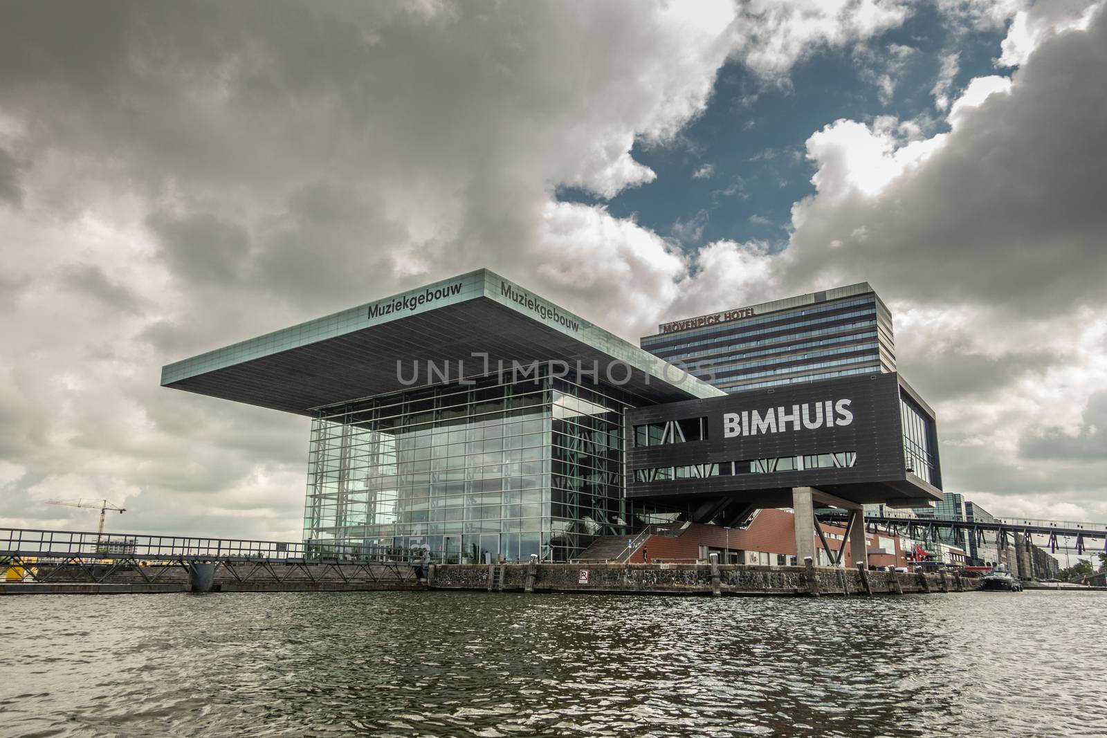 Amsterdam, the Netherlands - July 1, 2019: Modern cubic building housing the music theater and school with Jazz Bimhuis on platform in black water IJ river under havey rainy cloudscape.