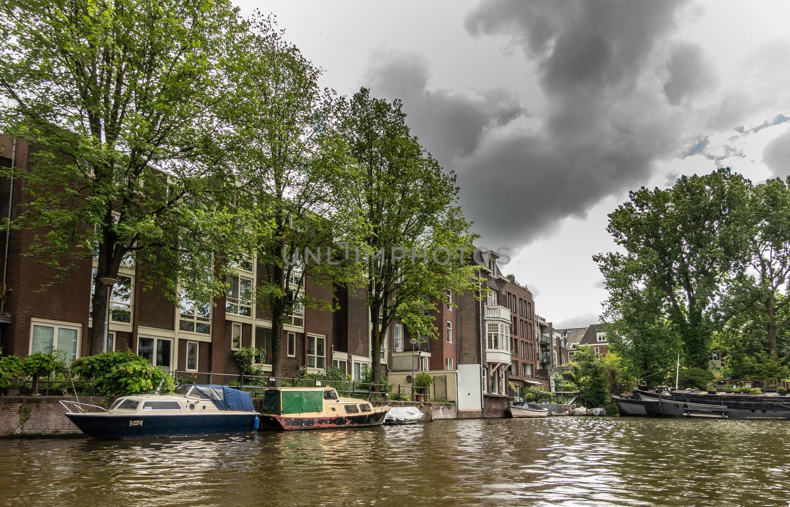 Amsterdam, the Netherlands - July 1, 2019: Peaceful canal boat with houseboats, green trees and apartment buildings under heavy cloudscape. Greenish water.