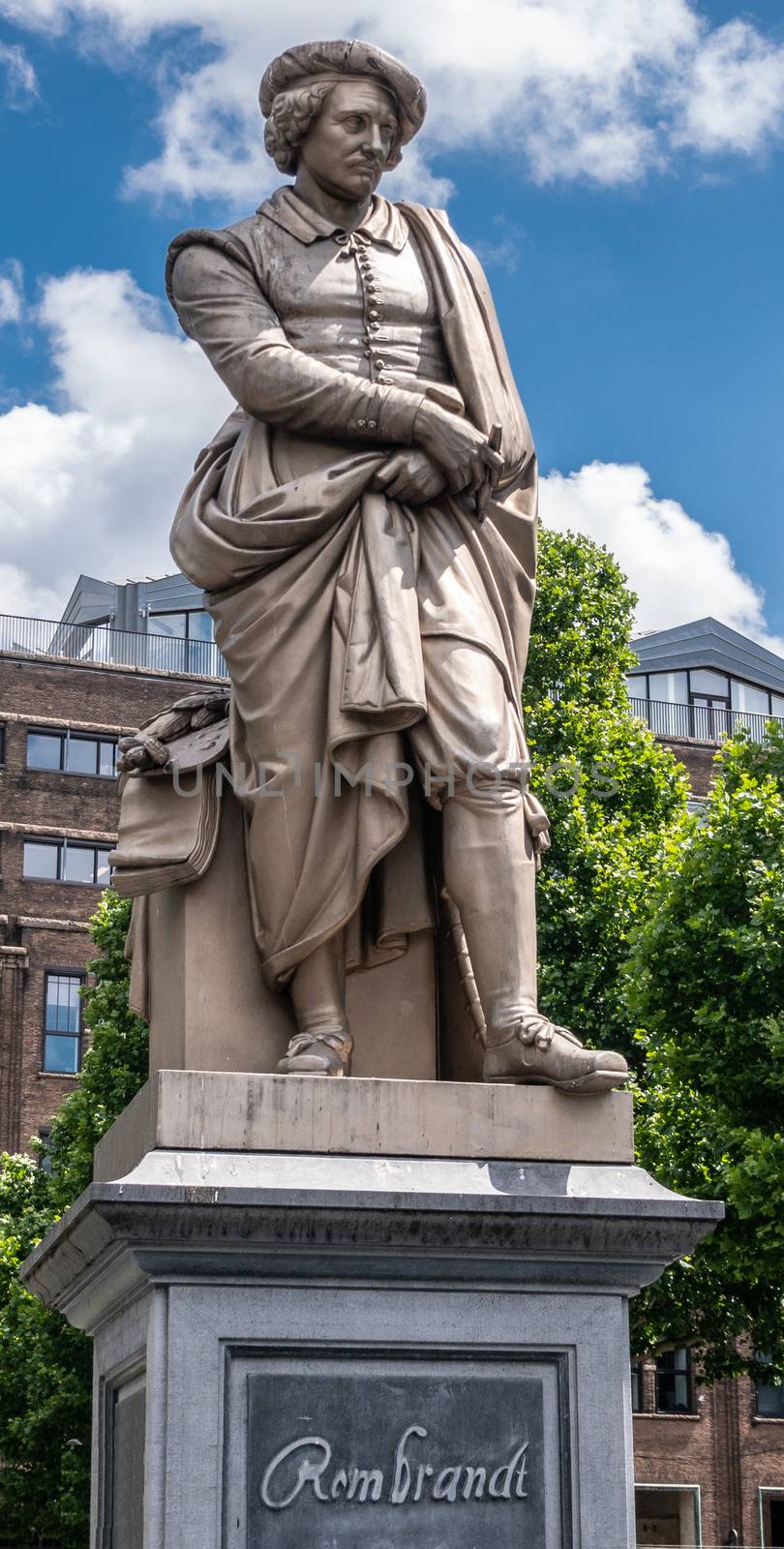 Amsterdam, the Netherlands - July 1, 2019: Beige full body of Rembrandt van Rijn on pedestal with his name statue on Rembrandtplein under blue sky with white clouds. tall buildings in back.