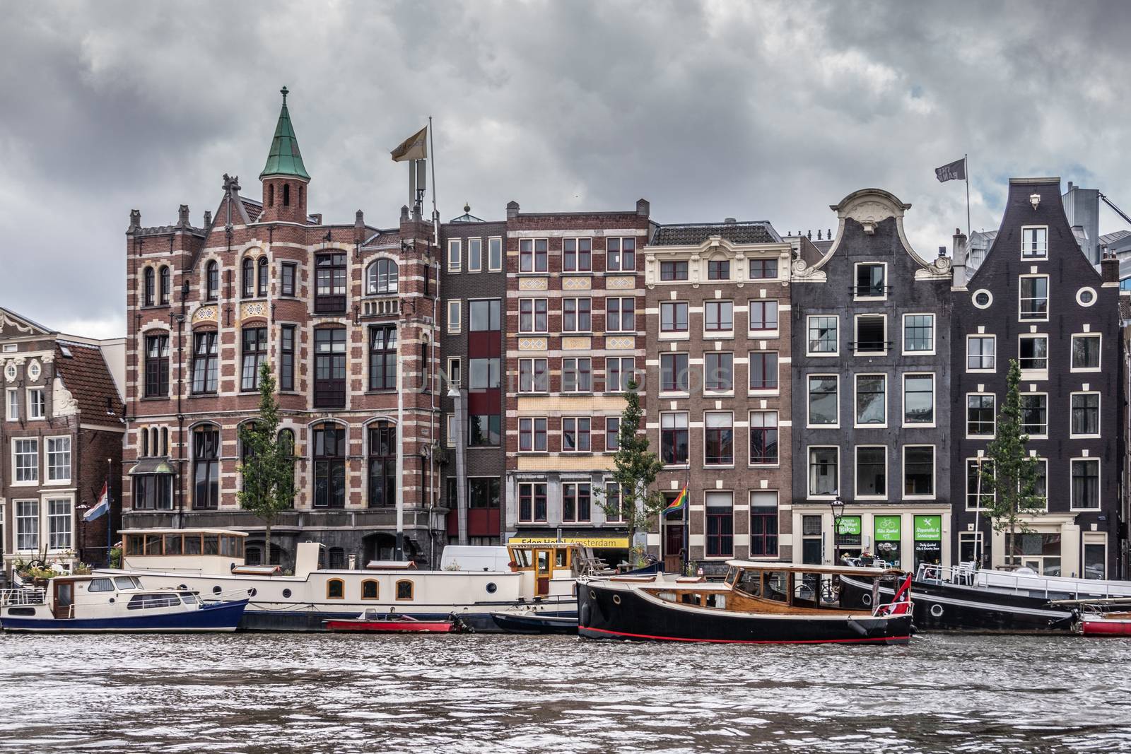Amsterdam, the Netherlands - July 1, 2019: Line of historic houses with gables along Amstel River downtown. Many houseboats and other vessels on dark river under heavy cloudscape.