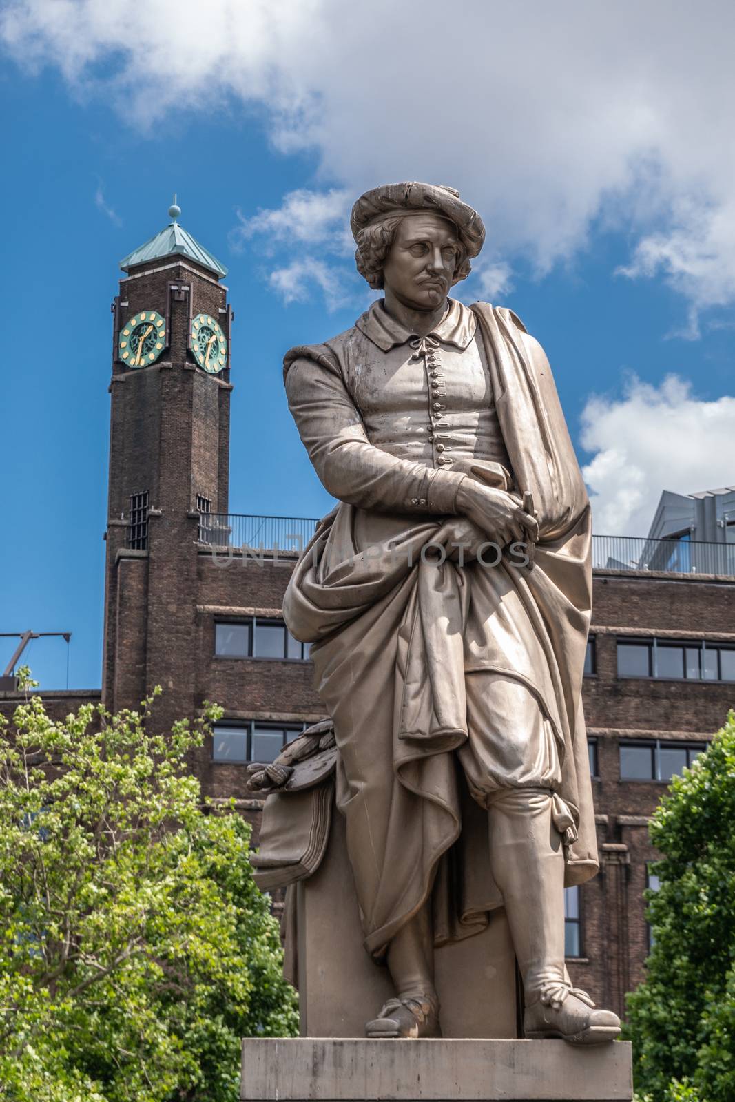 Amsterdam, the Netherlands - July 1, 2019: Beige full body of Rembrandt van Rijn statue on Rembrandtplein under blue sky with white clouds. Clock tower in brown stones in back.