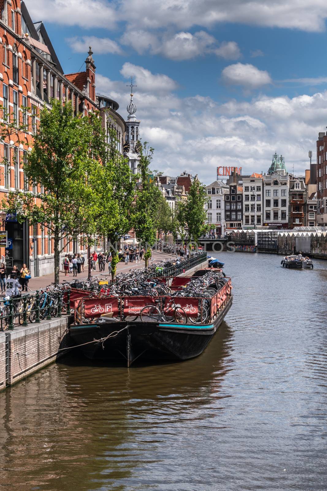 Amsterdam, the Netherlands - July 1, 2019: Dark flat barge is used as floating bike parking on Singel canal under blue-gray cloudscape. Munt tower spire. Glass houses of floating flower market on other side.