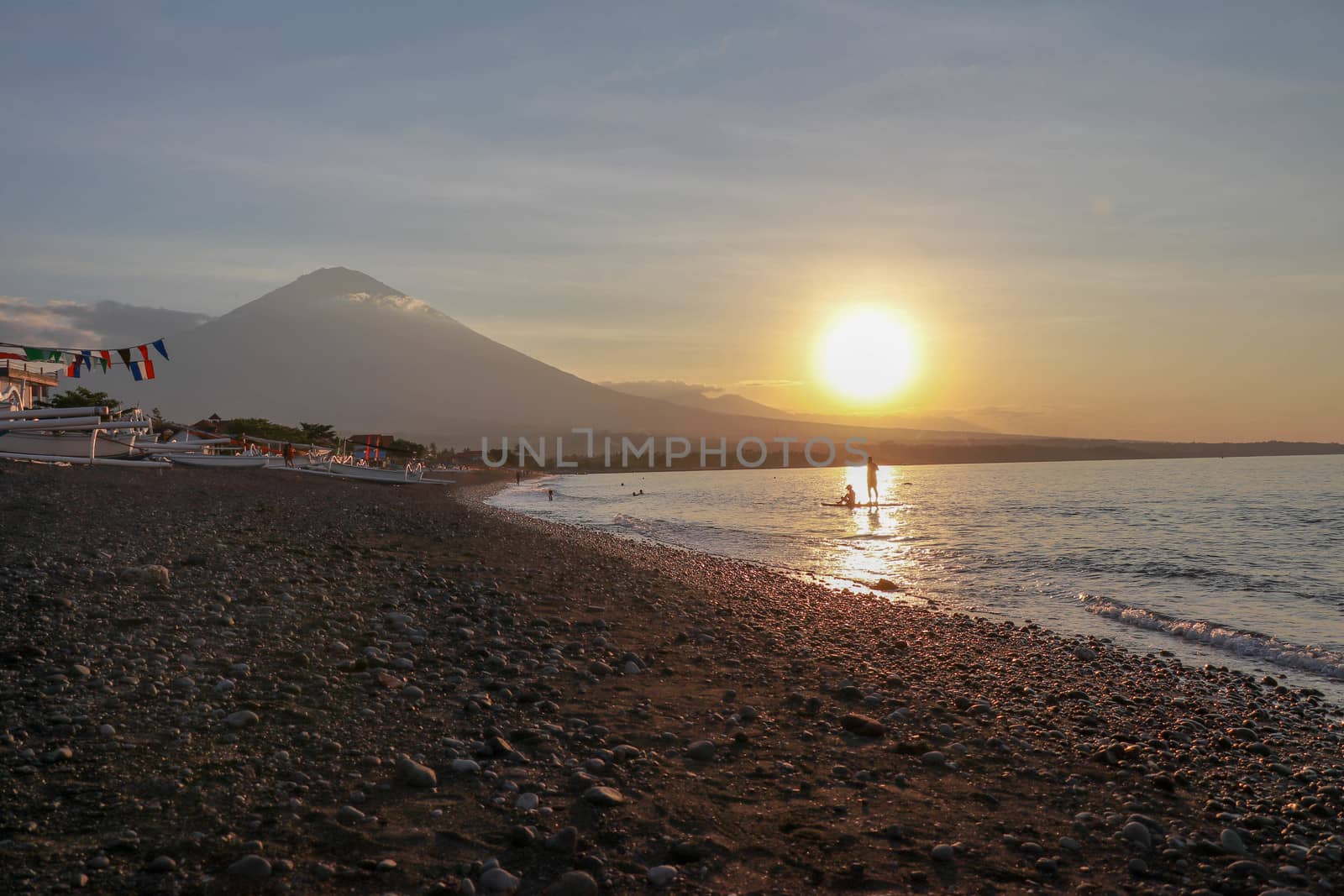 People enjoying surf and paddleboarding at sunset on a tropical island. Panorama with volcano and mountain range. Fishing boats parked on the beach.