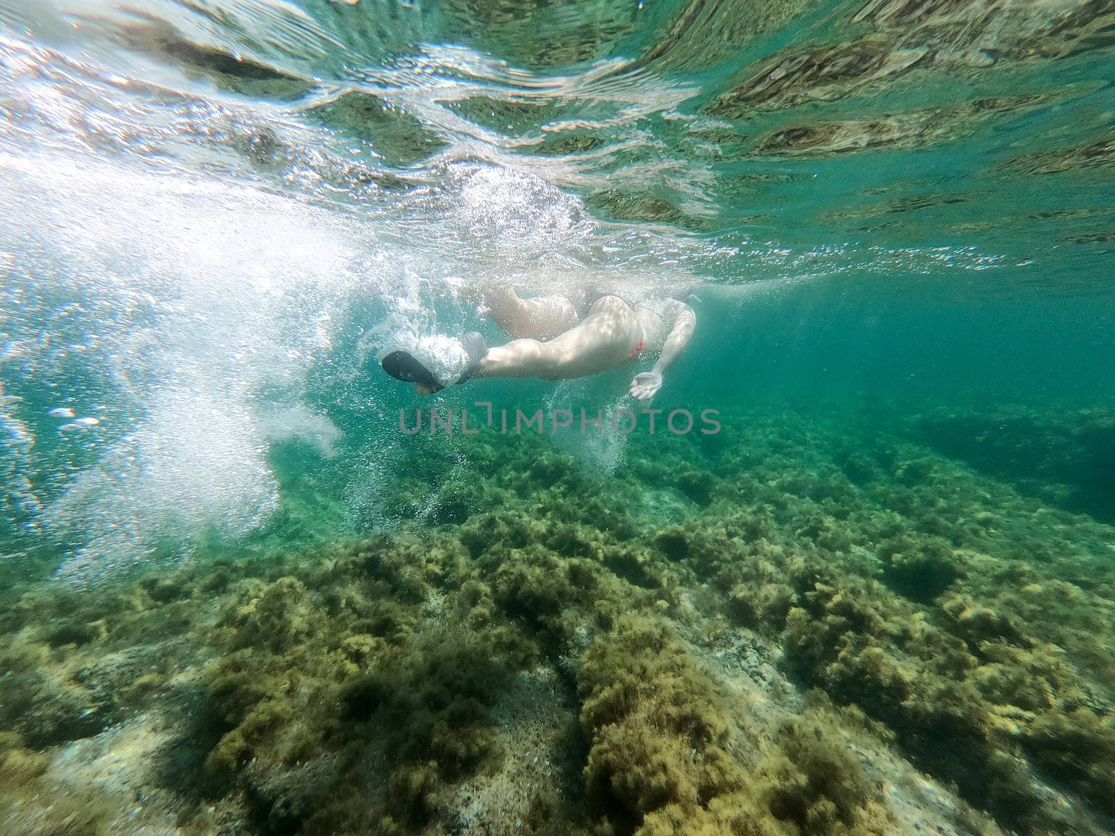 Young woman in bikini shooting underwater swims in the crystal clear water of the sea on rocks leaving a trail of bubbles with the reflection of her body by robbyfontanesi