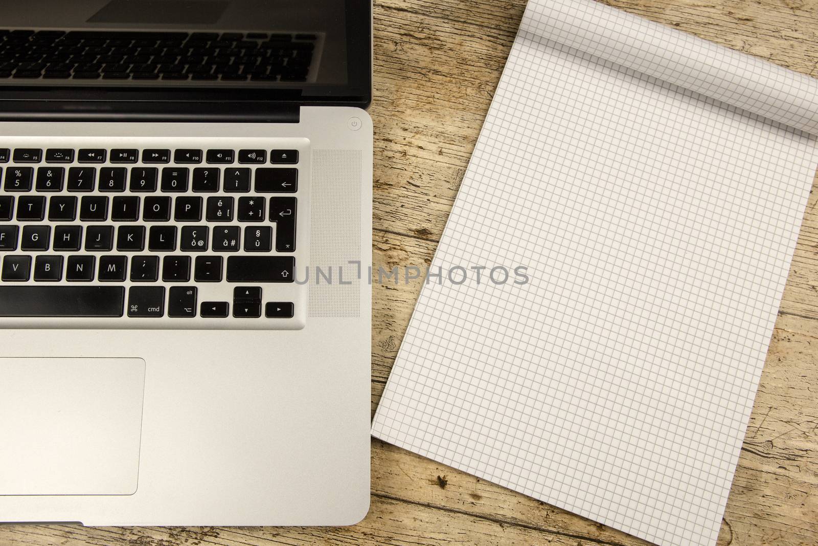 Contrast between old and new office work: shot from the top of an aluminum laptop on a wooden desk with an open checkered notebook with blank sheet