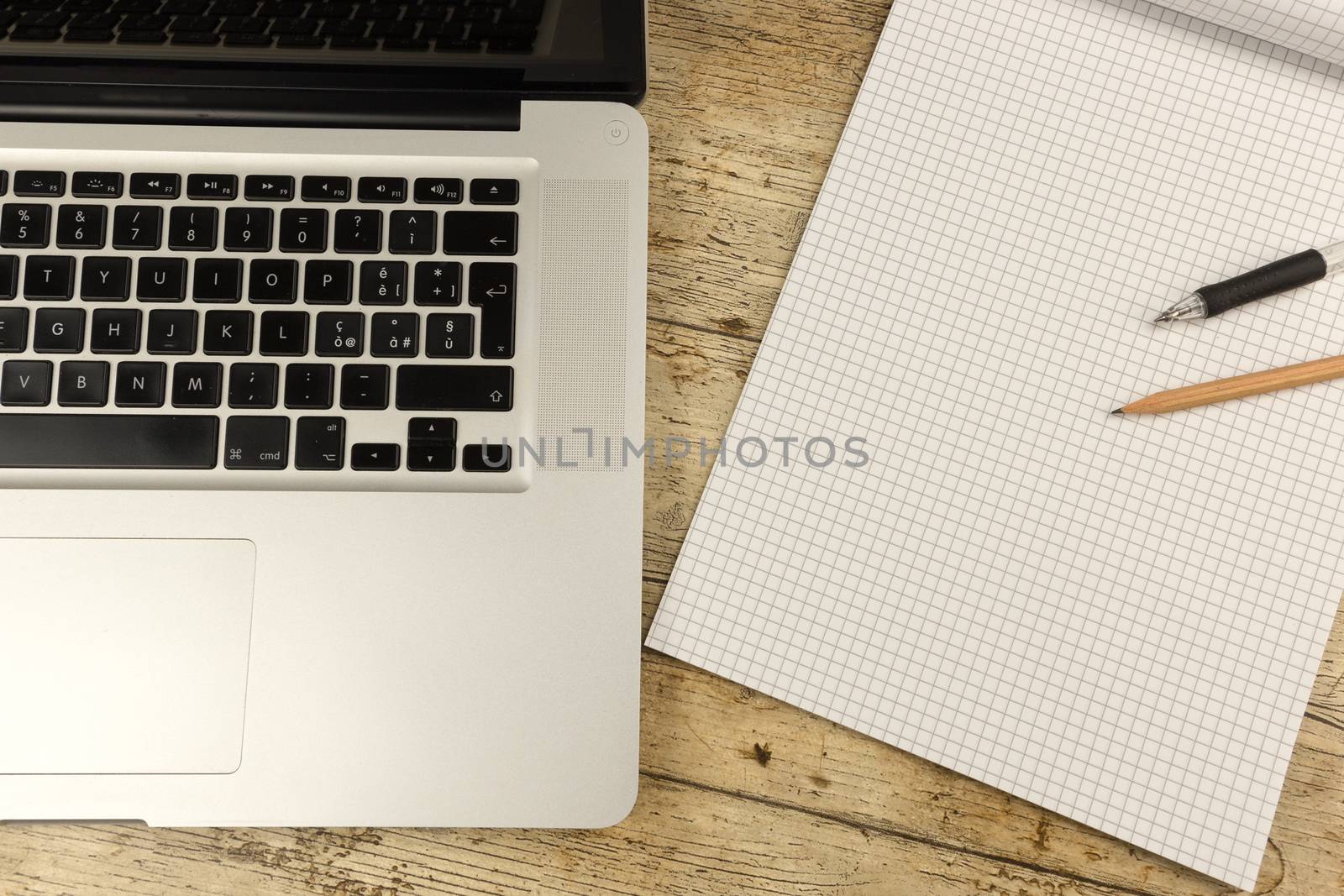 Contrast between old and new office work: shot from the top of an aluminum laptop on a wooden desk with an open checkered notebook, a pencil and a pen by robbyfontanesi