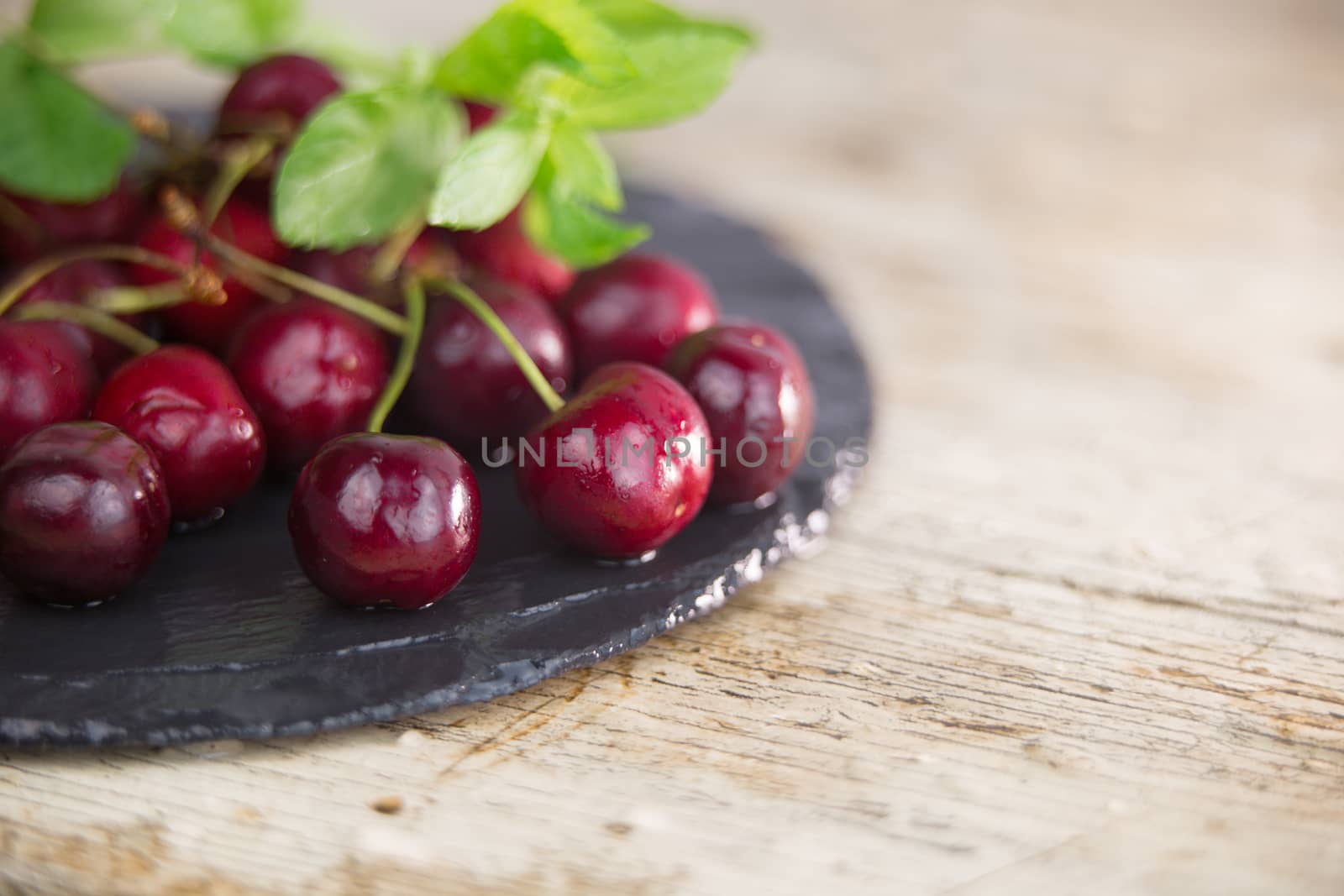 Fresh cherries on a black plate of wet slate with sprig of fresh green mint on light wooden table in selective focus for copy space by robbyfontanesi
