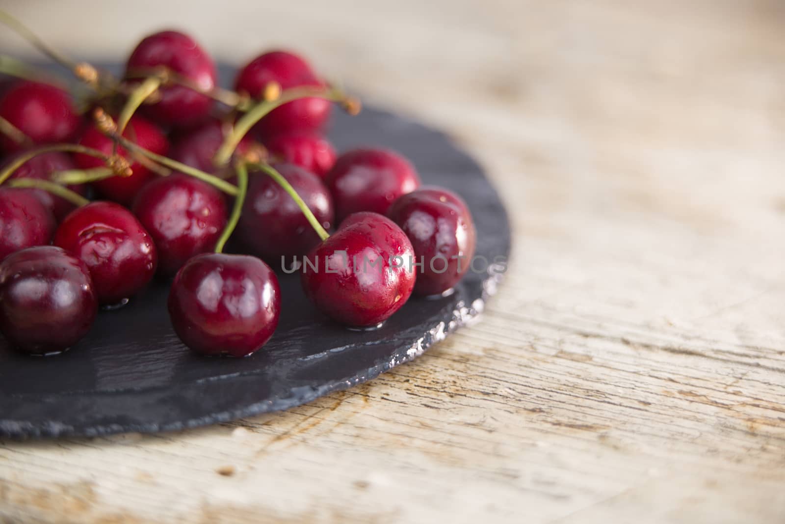 Fresh cherries on a black plate of wet slate on light wooden table in selective focus for copy space by robbyfontanesi