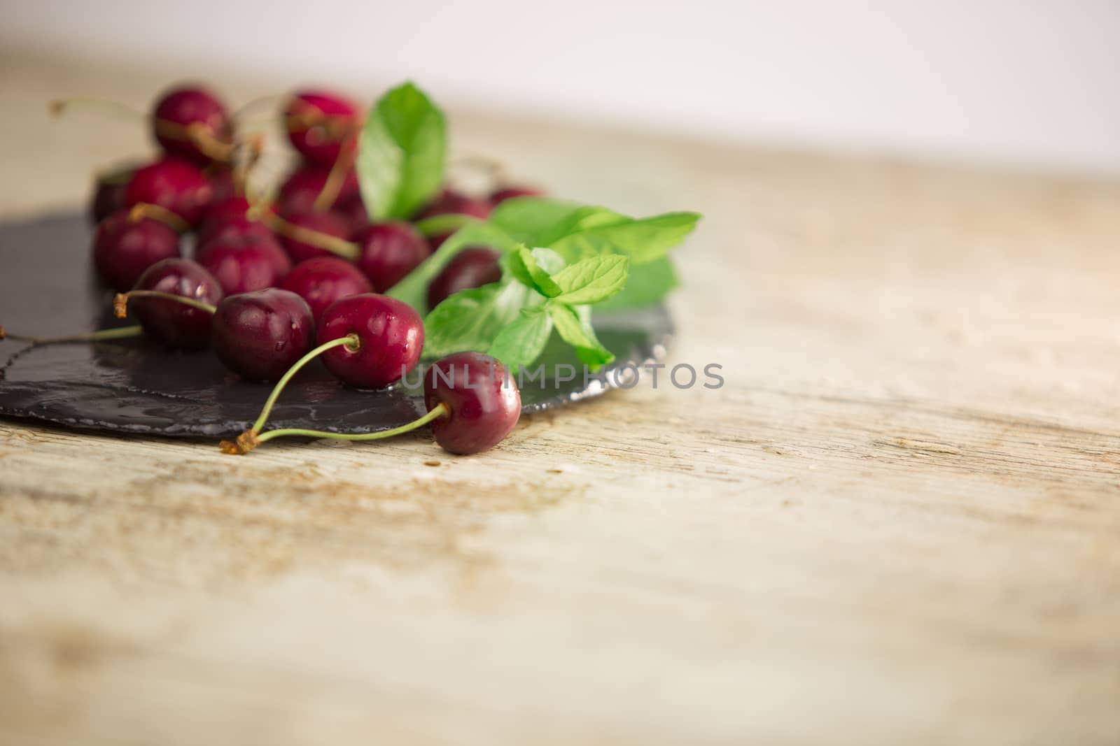 Fresh cherries on a black plate of wet slate with sprig of fresh green mint on light wooden table in selective focus for copy space